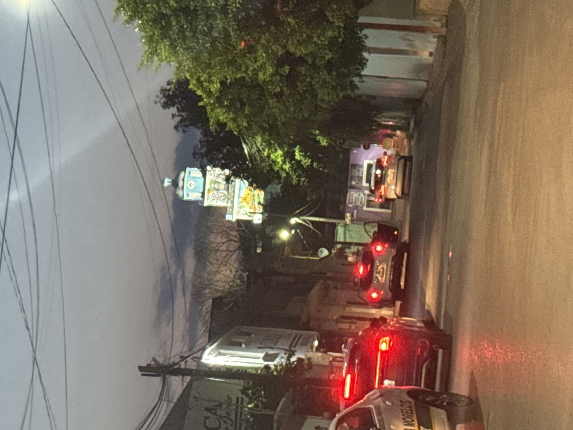 The image is a street scene in Xochitepec, Morelos, Mexico, at dusk or night. The photograph shows a line of cars driving on a street, their headlights and taillights illuminated. On the left side of the street, there is a tall structure with colorful decorations, possibly a church or a monument, lit up against the evening sky. There are trees and other buildings on both sides of the street. The road surface appears wet, reflecting the lights of the vehicles and the illuminated structure. Power lines are visible overhead.
