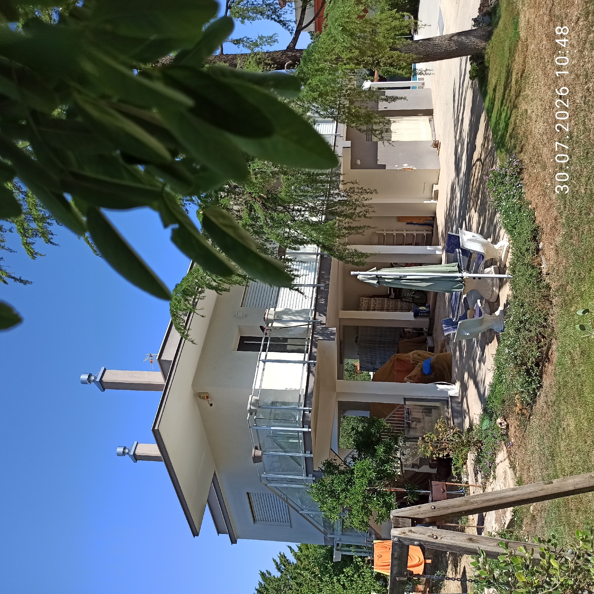The image depicts a sunny day in Terlizzi, Italy, in the backyard of a modern house. The house features a light-colored facade, multiple levels, and a balcony with glass railings. Two chimneys are visible on the roof. In the foreground, a wooden swing set stands on a grassy lawn. To the left, orange fabric is draped over a fence. An outdoor seating area with a table and chairs, covered by a green umbrella, is situated on a paved patio beneath the house. Lush green trees and foliage surround the property. The date and time stamp "30.07.2026 10:48" is visible in the lower right corner. There are no people present in the scene.