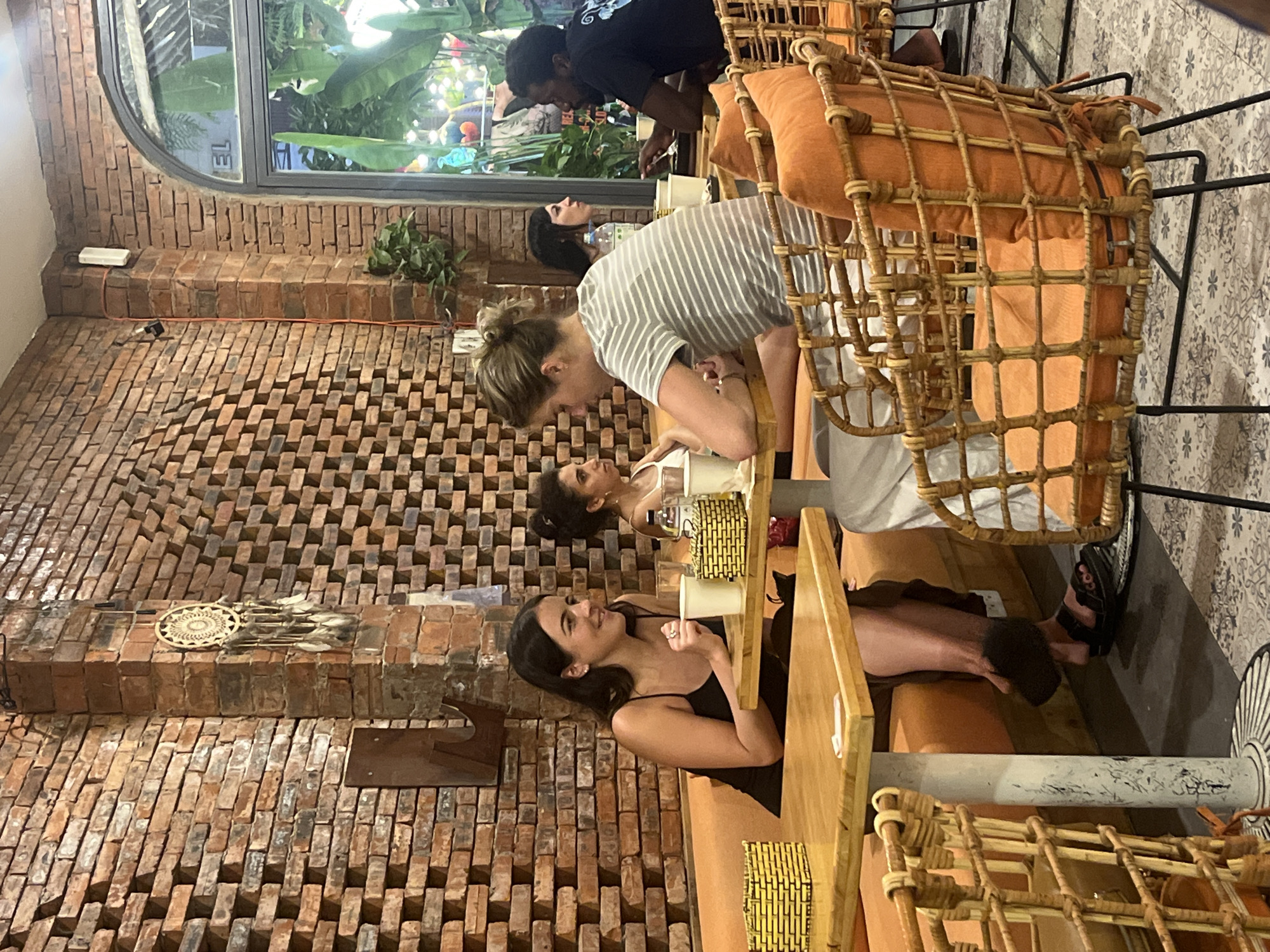 The image shows a group of people seated at tables in a dimly lit, indoor establishment with exposed brick walls. The people are engaged in conversation and appear to be dining or socializing. The setting suggests a restaurant or cafe, possibly with an outdoor area visible through a large window in the background, where lush green plants and some decorative lights are visible. The furniture includes chairs with woven rattan frames and orange cushions.
One woman, wearing a black spaghetti strap dress, is seated at the foreground table, smiling and looking to her left. Another woman, in a striped t-shirt, leans forward across the table, seemingly interacting with someone seated further away. A man in a dark t-shirt is visible in the background, leaning over a table. The overall mood seems relaxed and social.
Notable details include the intricate brickwork of the walls, decorative elements like a circular wall hanging with feathers, and the warm orange tones of the cushions. The lighting suggests it might be evening or a dimly lit indoor space. No visible text is discernible in the image. The location is identified as Đà Nẵng, Vietnam, which aligns with the tropical foliage seen through the window.