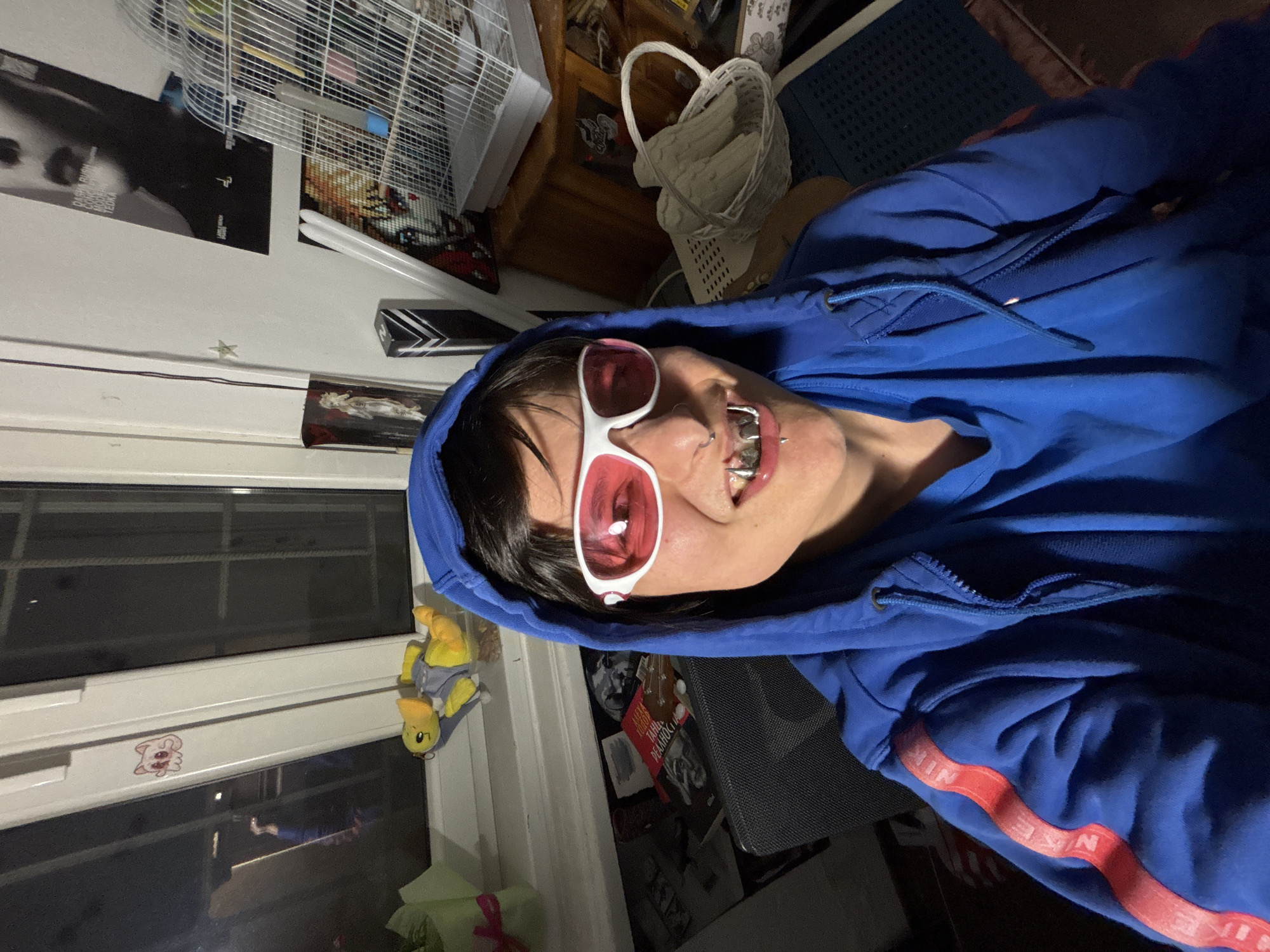 A person wearing a blue hoodie and white-framed sunglasses with pink lenses is posing for a selfie. The person has silver-colored dental grills on their teeth. The background includes a window with a stuffed duck hanging on it, a birdcage, a white wicker basket, and posters on the wall. The location cannot be determined to be Sofia, Bulgaria from the image.