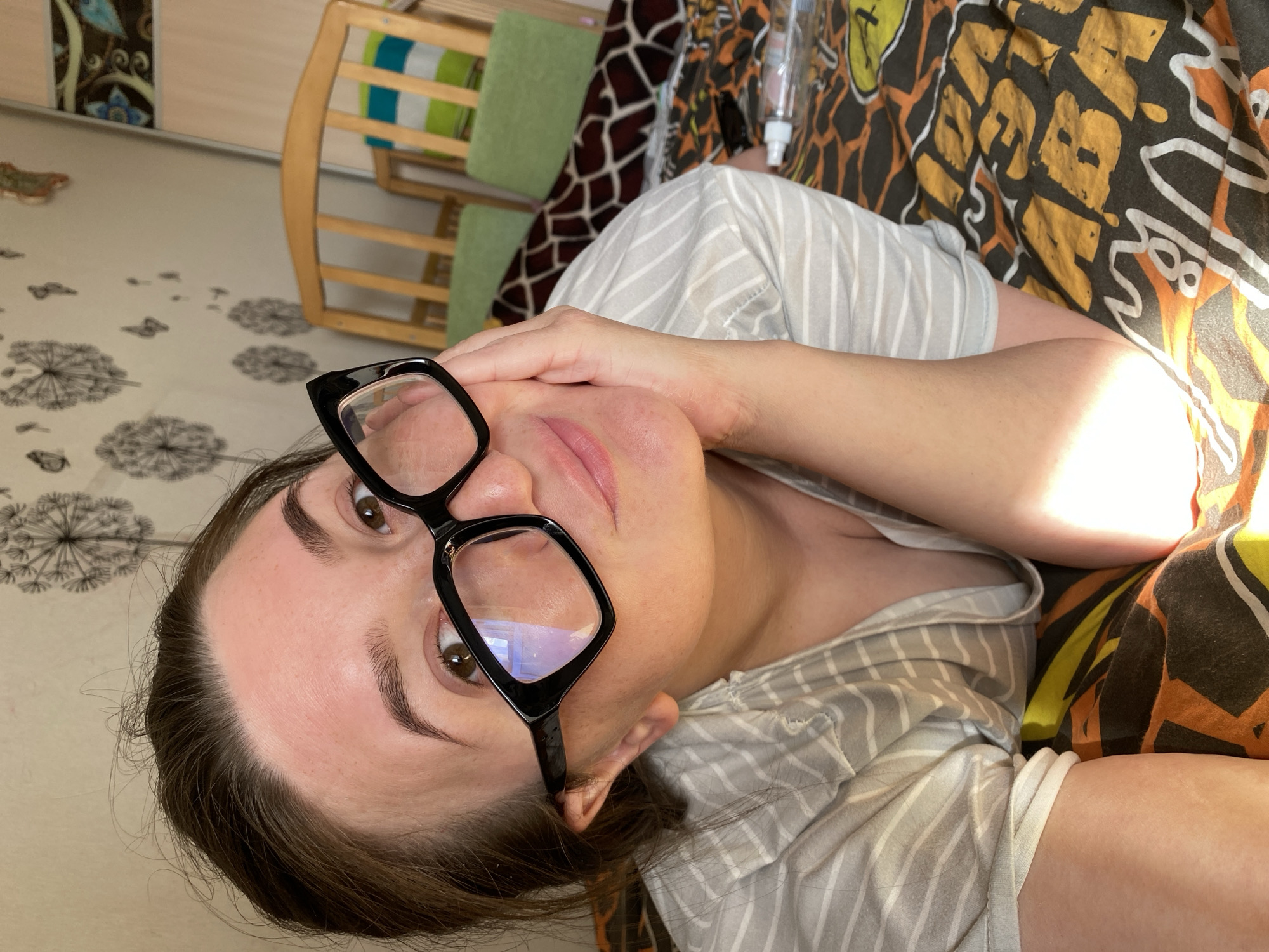 A woman with brown hair and wearing black framed glasses is looking at the camera. She is wearing a gray and white striped shirt and lying on a bed with a black, yellow, and brown patterned blanket. A baby crib with a green cushion is visible in the background. The location is likely an indoor setting in Chaikovsky, Russia, indicated by the user prompt. The woman is resting her right hand on her face. A transparent plastic bottle is visible near the head of the bed. The floor has a dandelion patterned design.