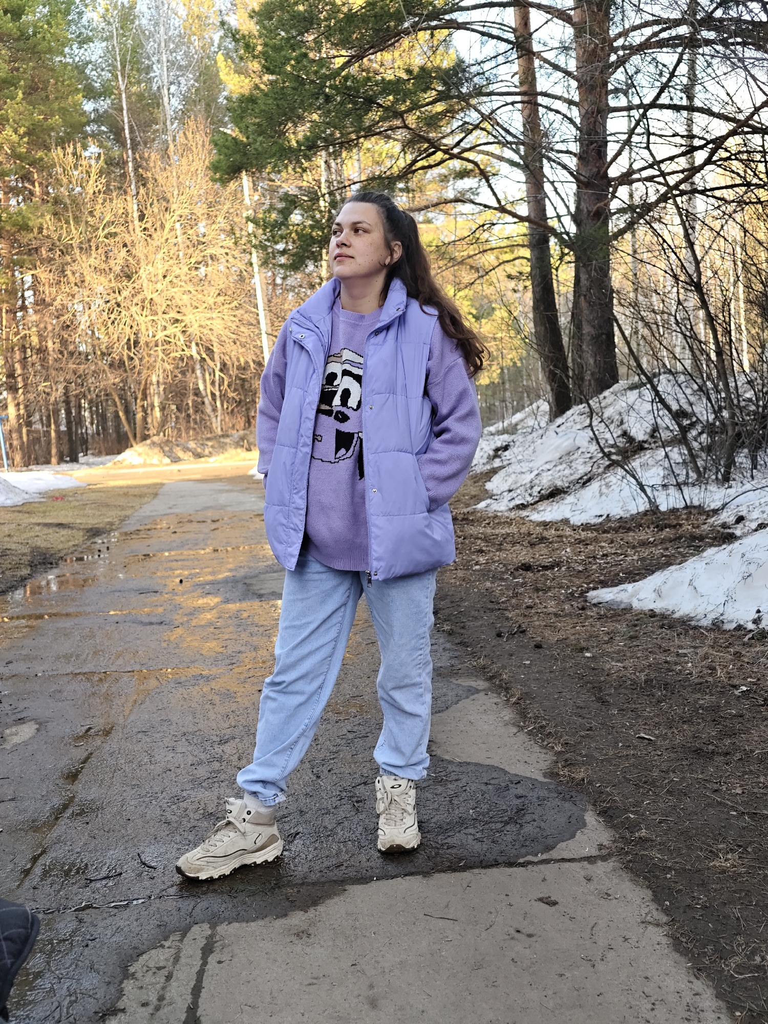 A woman stands on a wet, cracked asphalt road within a park setting. She wears a lavender sweater with a graphic design, light blue jeans, a lavender puffy vest, and off-white sneakers. Her dark hair is pulled back in a ponytail. The area around her is a mix of bare trees, some snow patches on the ground, and damp earth. The background suggests a wooded area with a park-like setting. This is most likely a park in the city of Chaikovsky, Russia.