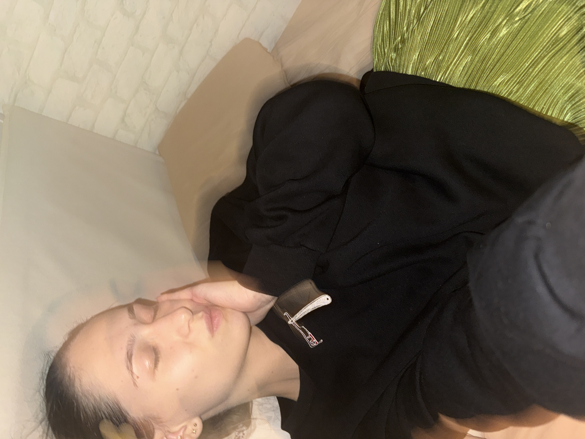 A young woman with dark hair pulled back is lying down, her eyes closed and her right hand resting on her cheek. She is wearing a black long-sleeved shirt with a brooch of a straight razor on the left side of her chest and a green pleated skirt. The background consists of a beige pillow and a wall with a brick-patterned wallpaper. The lighting is dim, casting shadows on the wall. The scene appears to be indoors, possibly in a residential setting in Sofia, Bulgaria.