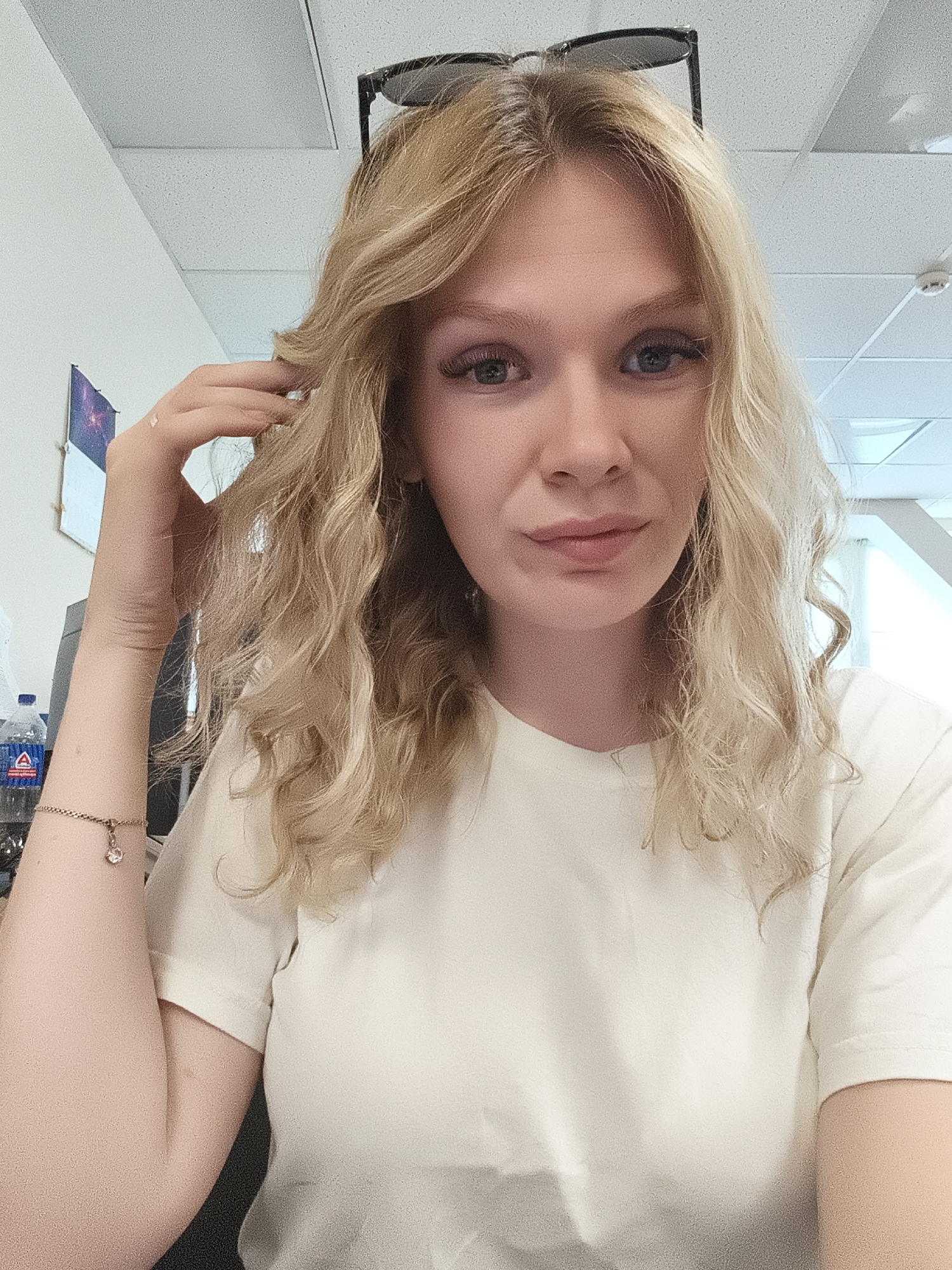 A young Caucasian female with blonde hair is photographed from the chest up, looking directly at the camera. She is wearing a light-colored t-shirt and has sunglasses perched on top of her head. Her right hand is raised and her fingers are touching her hair. She is wearing a bracelet on her left wrist with a charm. The background appears to be an indoor office setting with white walls, a drop ceiling, and fluorescent lighting. There are also some filing cabinets and a bottle of water visible in the background. The overall lighting is bright and even, suggesting the photo was taken during the day.