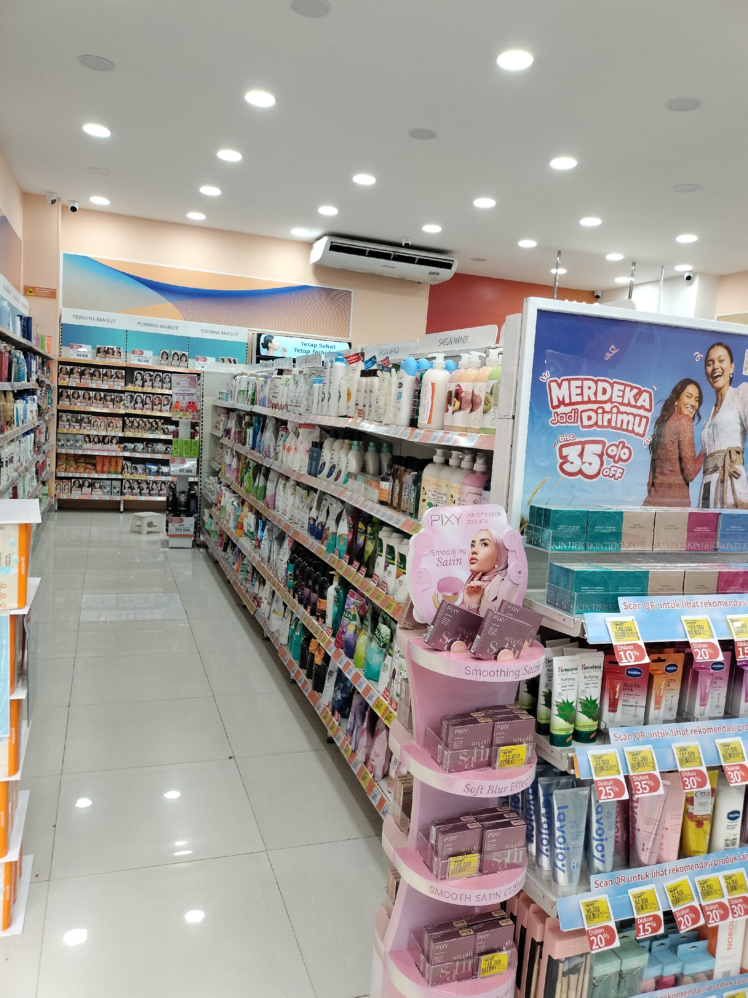 The image depicts the interior of a well-lit retail store, likely a pharmacy or a personal care product store, located in Langsa, Indonesia. The scene is dominated by shelves stocked with a variety of products, including shampoos, conditioners, soaps, lotions, and makeup. In the foreground, a display stand showcases PIXY brand makeup products, featuring prominent signage for "Smoothing Satin Cushion" and "Soft Blur Effect." Further back, shelves are organized by product categories, with large promotional posters visible on the walls. One poster displays "MERDEKA Jadi Dirimu" with a "35% OFF" discount and images of two women. The flooring is tiled and reflects the bright overhead lighting. Security cameras are visible on the ceiling, indicating a standard retail security setup. The overall impression is that of a clean, organized, and modern retail environment offering a wide range of consumer goods.