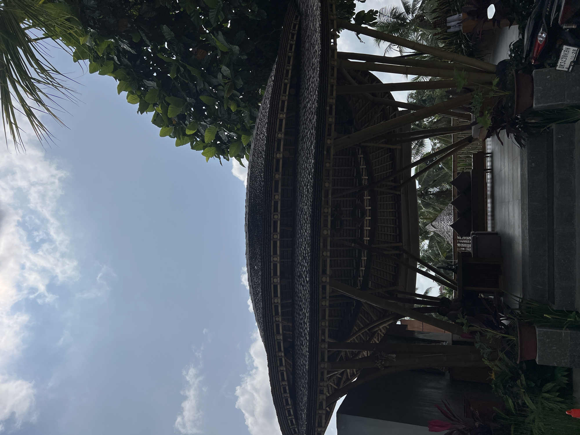 The image shows a bamboo structure in what appears to be a tropical setting, likely in the Ubud District of Indonesia. The structure has a multi-tiered, curving roof made of dark material resembling thatch or wooden shingles. It is supported by a bamboo frame, and the interior construction is visible. Adjacent to the structure are planters with greenery, a set of steps made of dark stone, and a parked scooter. Part of the foliage of a tree and the sky with light clouds are visible.