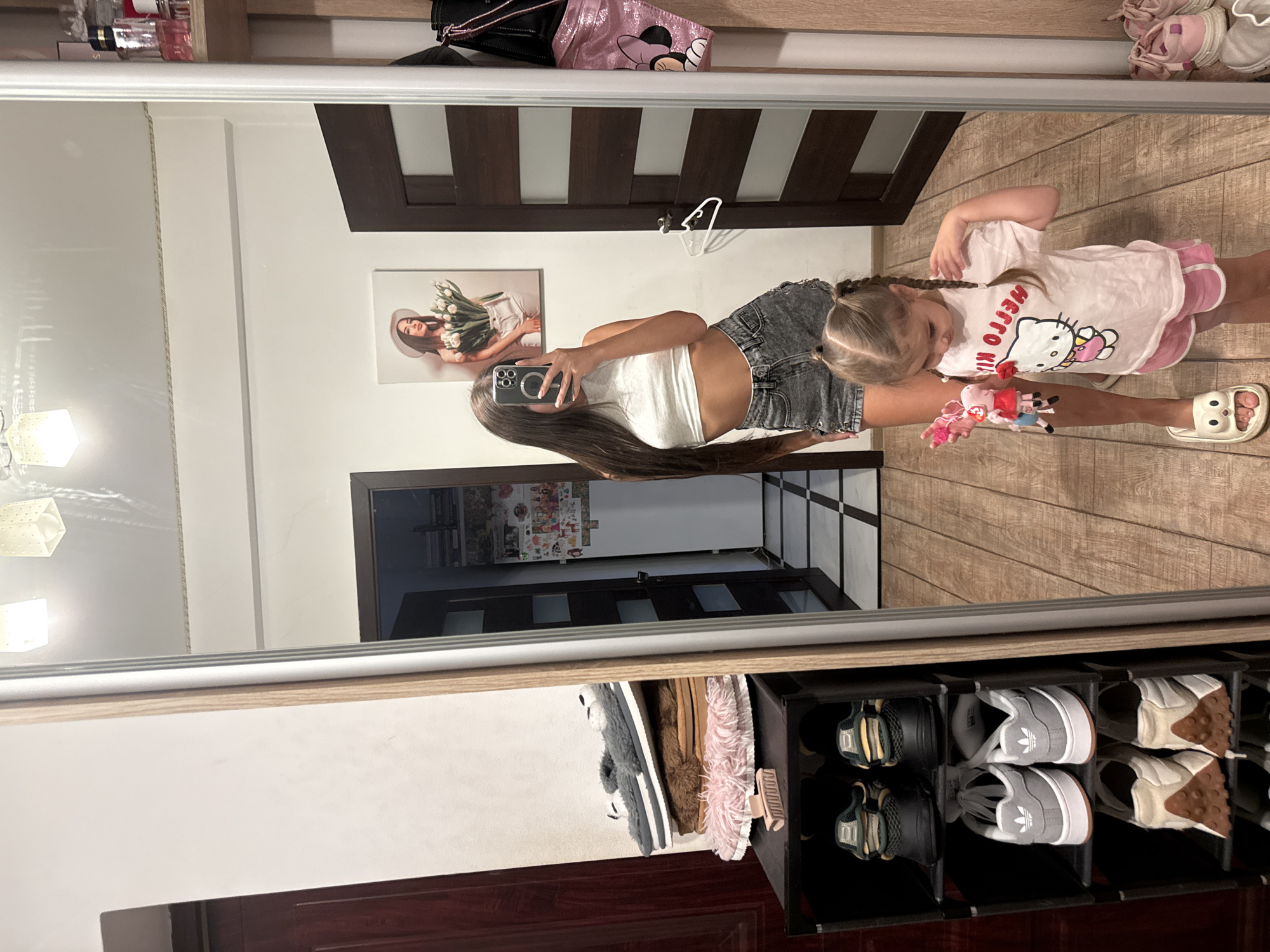 The image shows a reflection in a full-length mirror of a young woman taking a selfie. She is wearing a white crop top and gray denim shorts. A young child is standing next to her, wearing a pink t-shirt with a Hello Kitty graphic and pink shorts. The child is holding a small stuffed toy.
The background of the reflection reveals an interior setting, likely a home. There is a shoe rack with several pairs of shoes, a shelf with bottles of perfume, and a painting on the wall of a woman holding flowers. There are also two doors visible. The floor appears to be tiled in some areas and wooden in others.
There is no information in the image or its crops that suggests the location is in Chișinău, Moldova.