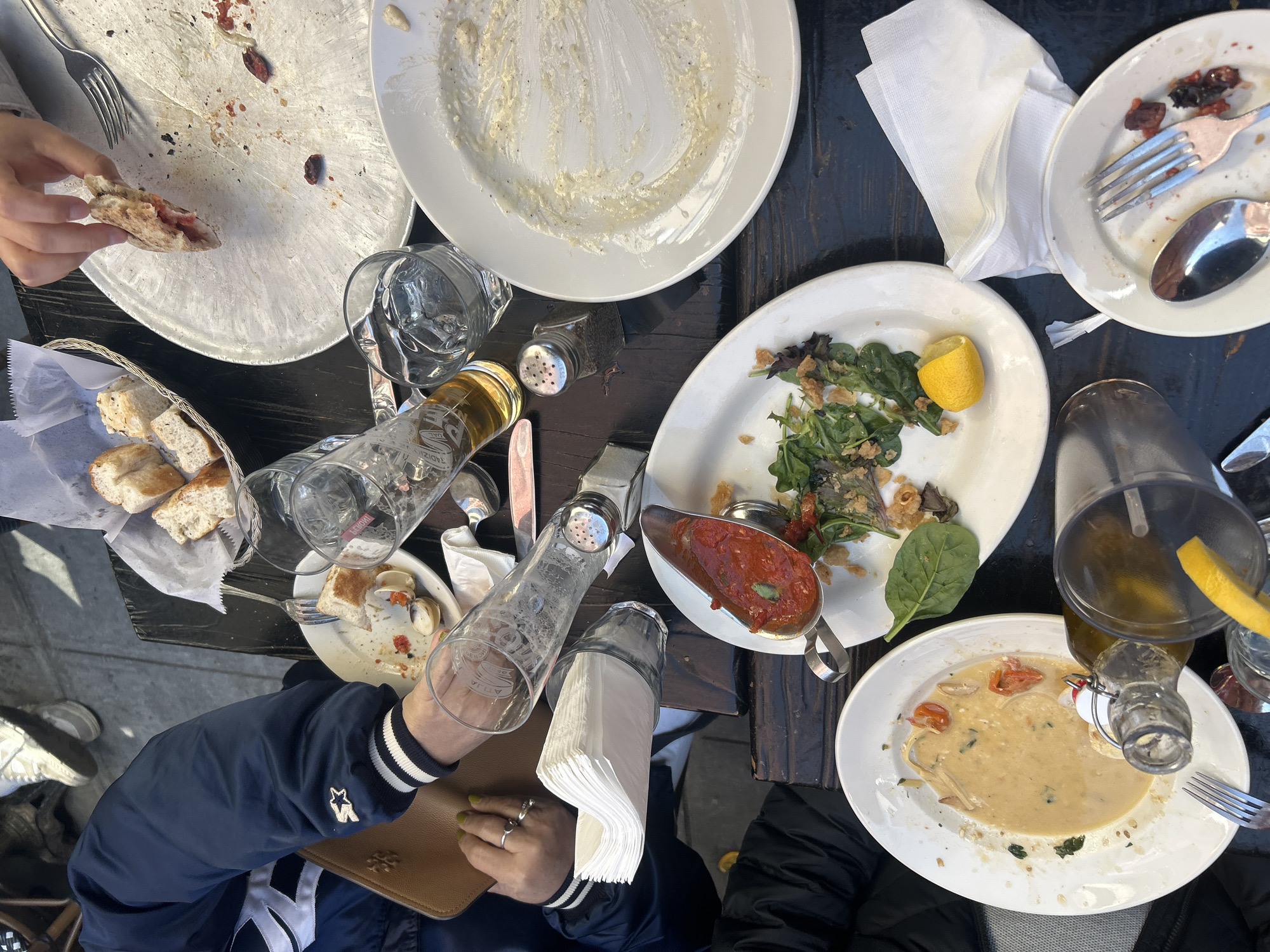 The image captures an aerial view of a table laden with the remnants of a meal, set in what appears to be an outdoor dining area. Several empty plates, some with residual food, suggest a shared dining experience. There are also glasses, bottles, silverware, and a basket of bread. Two people are partially visible at the bottom of the frame; one is wearing a navy blue jacket with a star and V logo and holding a brown leather purse, and the other is in a black jacket. A hand holding a piece of pizza is visible in the upper left. The environment seems to be urban, potentially in the city of New York, United States of America, considering the style of dining and the general atmosphere. There are no immediately identifiable landmarks that would confirm the location.
