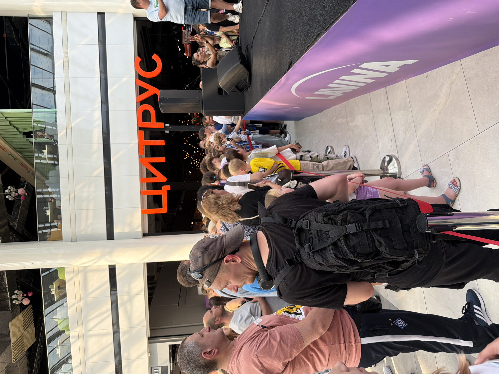 This image shows a crowd of people gathered in front of a stage in what appears to be an indoor shopping mall in Kyiv, Ukraine. The word "ЦИТРУС" (Citrus) is prominently displayed in large orange letters above the stage, suggesting this is an event or promotion for a store or brand with that name. Several people are in line, and some are looking towards the stage, where a woman is seen taking a picture with her phone. The event seems to be outdoors within a large, modern building with multiple levels visible in the background. The overall scene depicts a public gathering, possibly for entertainment or a product launch.
