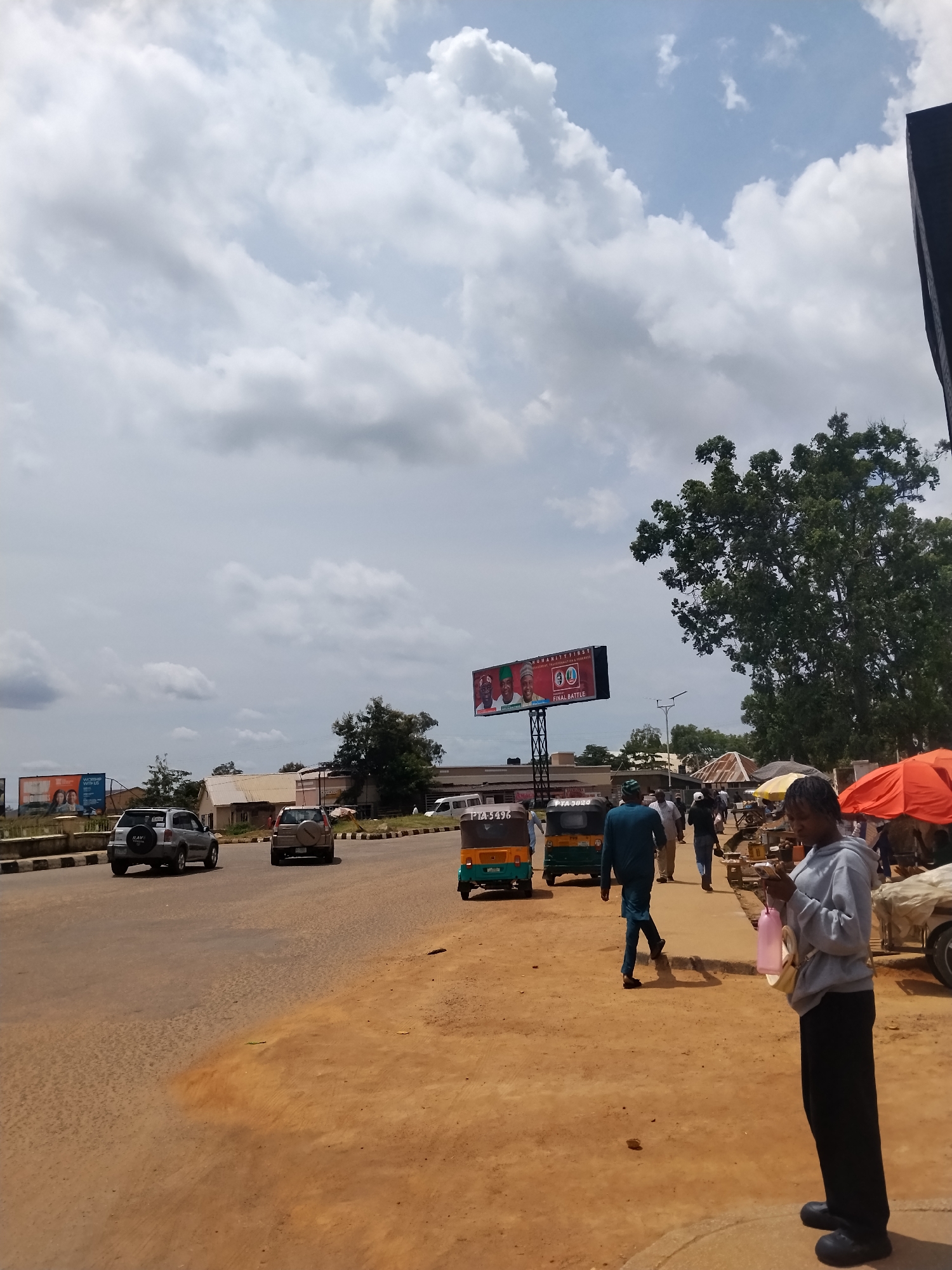 This image captures a street scene in Jos, Nigeria, under a cloudy sky. The road is paved, with a dusty, reddish-brown shoulder on the right side where a few people are present. In the foreground, a young woman in a grey hooded sweatshirt and black pants stands facing right, holding a pink bottle and a light-colored bag. Behind her, several other individuals walk along the sidewalk, some under temporary awnings.
A billboard is prominently displayed in the mid-ground, featuring images of three men in political attire, with text that appears to read "HUMANITY FIRST" and "FINAL BATTLE." Beside the billboard, there are several three-wheeled vehicles (Keke NAPEPs) and cars, including a silver SUV. Further down the road, more vehicles are visible, and buildings line the street. To the far right, an orange umbrella shades a market stall. The overall impression is of a bustling urban environment on a partly cloudy day.