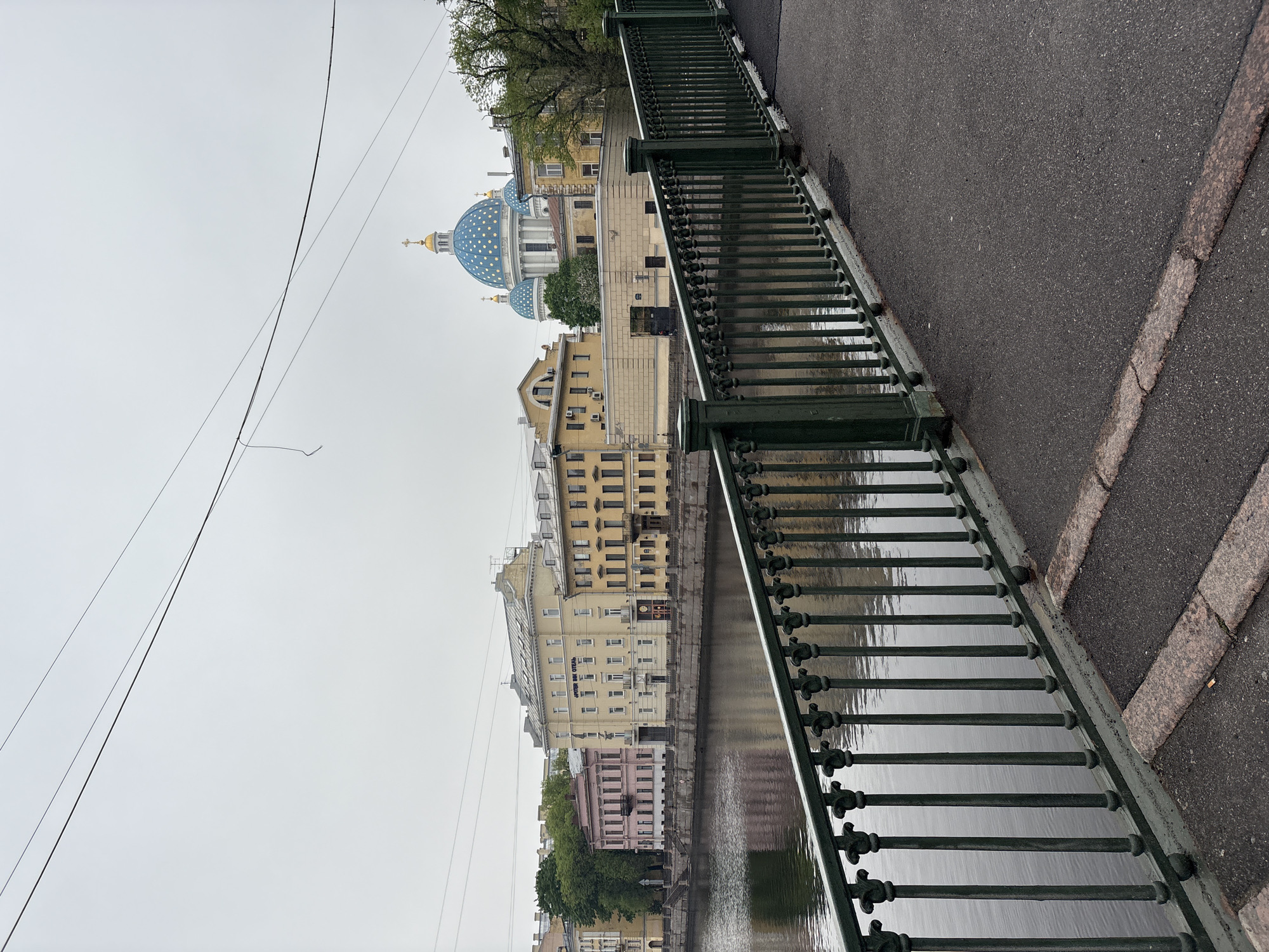 A photograph shows a bridge in Saint Petersburg, Russia, with a view of the Smolny Cathedral in the background. The green metal railing of the bridge is in the foreground, casting a reflection onto the water below. Adjacent to the railing is a paved sidewalk. Across the canal, several buildings of varying heights and colors, including light yellow and pink, line the embankment. The blue and gold domes of the Smolny Cathedral, adorned with star-like patterns, stand prominently against the overcast sky. Above the buildings, power lines are visible stretching across the sky. The weather appears overcast.