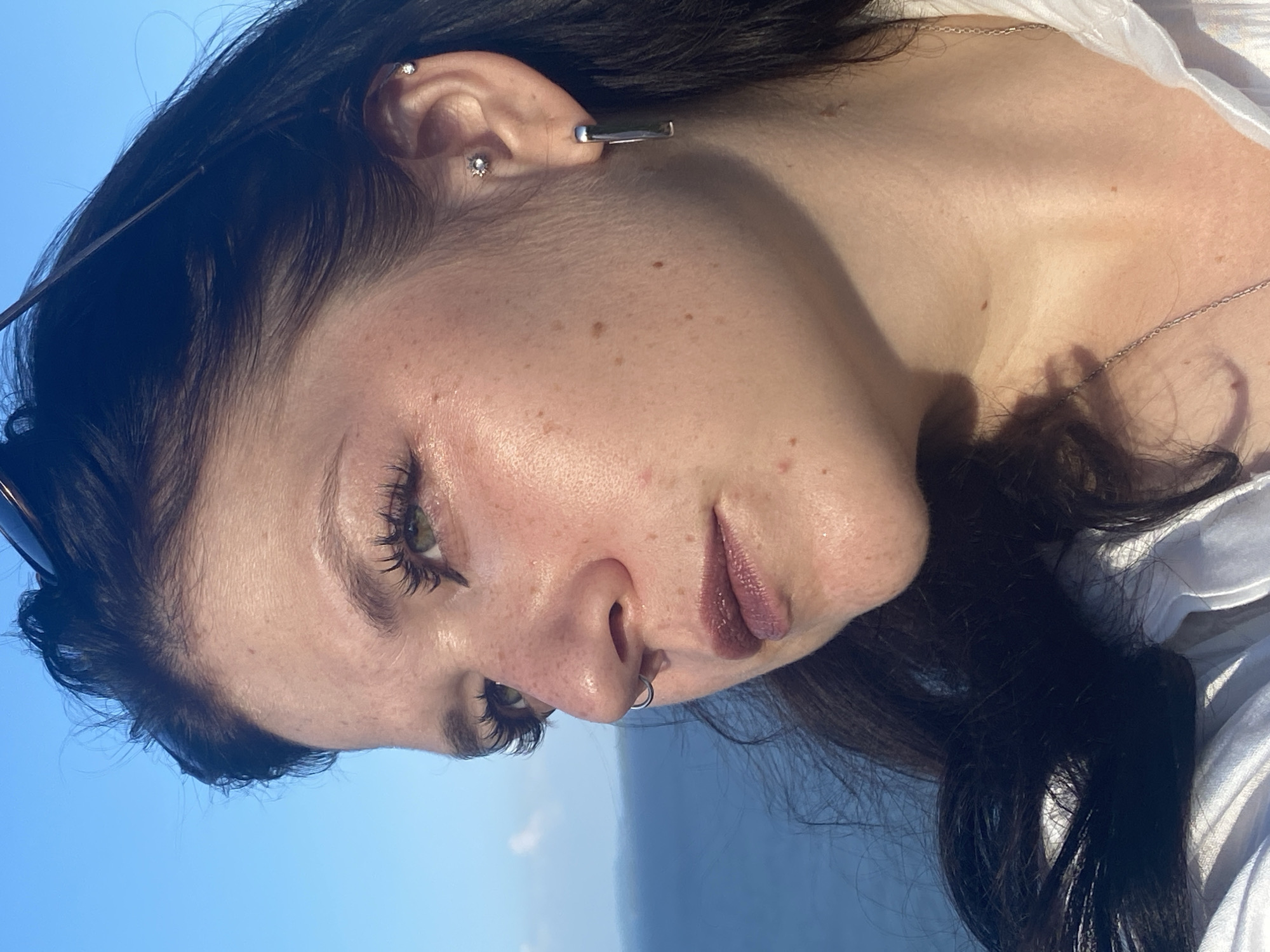 A close-up, profile shot captures a young woman with dark hair and light skin, her face turned to the left, gazing outwards. The background is a clear blue sky and a distant, hazy blue body of water, suggesting an outdoor setting, possibly a coast or a large lake. The woman has light green eyes, defined eyebrows, and prominent eyelashes. She is wearing a silver hoop earring in her nose and two stud earrings in her left ear, one higher on the ear and another lower. A large, rectangular silver earring hangs from her earlobe. Her lips are painted with a dark berry lipstick. She is wearing a white collared shirt and a delicate silver chain necklace. Her hair is dark brown and long, with some strands falling across her face and shoulder. The lighting suggests it is daytime, with the sun casting a warm glow on her skin, highlighting freckles and subtle signs of aging. Sunglasses are perched on top of her head. The image does not contain any information or context that would place it in Kostovaty, Russia.