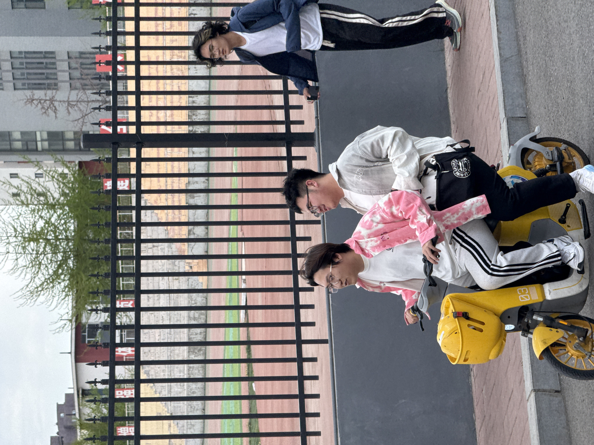 Three young people are outdoors in what appears to be an urban or campus setting in Zhongtun, China. The image is oriented sideways, with the ground appearing on the right.
In the foreground, a person with short dark hair and glasses is seated on a bright yellow shared e-bike or scooter, which has "03" marked on its front. This person wears a pink and white tie-dye jacket over a white t-shirt and light gray sweatpants with black stripes, along with white sneakers. Standing beside the bike is another person, also with short dark hair and glasses, wearing a light-colored jacket over a white t-shirt and dark pants. A black shoulder bag with a white circular logo, prominently displaying "LI-NING," hangs from their side. Further back, behind a black metal fence, a third person stands. This individual has longer, curly dark hair, wears glasses, a dark blue jacket over a white t-shirt, dark pants with white stripes, and sneakers. They are holding a small dark object in their hand.
The activity shows the individuals relaxing or socializing. The person on the bike appears to be stationary, while the other two stand nearby, looking generally towards the right side of the frame. Their expressions are neutral and casual.
Behind the fence, a sports track (brown) and a green field are visible, suggesting proximity to a sports facility. In the far background, buildings can be seen under an overcast sky, indicating it is daytime with soft light. Faint red banners in the distance behind the fence show some Chinese characters, including "禁止" (Jìnzhǐ - Prohibited/Forbidden) on one. The metal fence itself features decorative finials at the top of its vertical bars. The paved area where the people and bike are situated has sections of red brick and gray concrete.