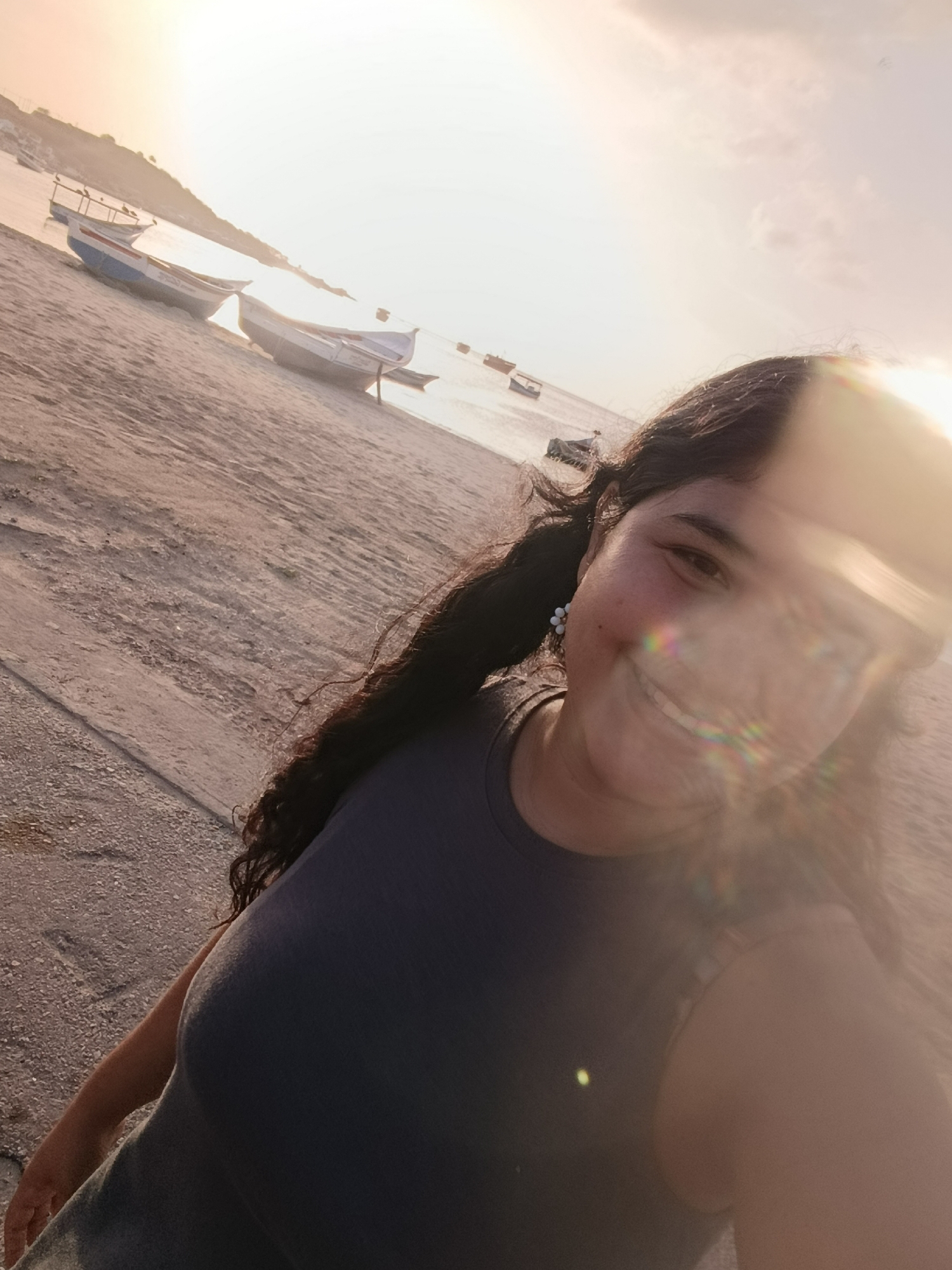 In Juan Griego, Venezuela, a young woman with long, dark, curly hair is captured in a selfie at a beach during sunset. The sun's glare creates lens flare across the image, obscuring parts of her face and body. She is wearing a dark blue tank top and has small earrings. Her hair is partially obscuring her face. The background shows a sandy beach with several small, traditional fishing boats pulled up onto the shore. The water appears calm, reflecting the soft, golden light of the setting sun, and a gentle slope of land is visible in the distance. The overall atmosphere is peaceful and serene.