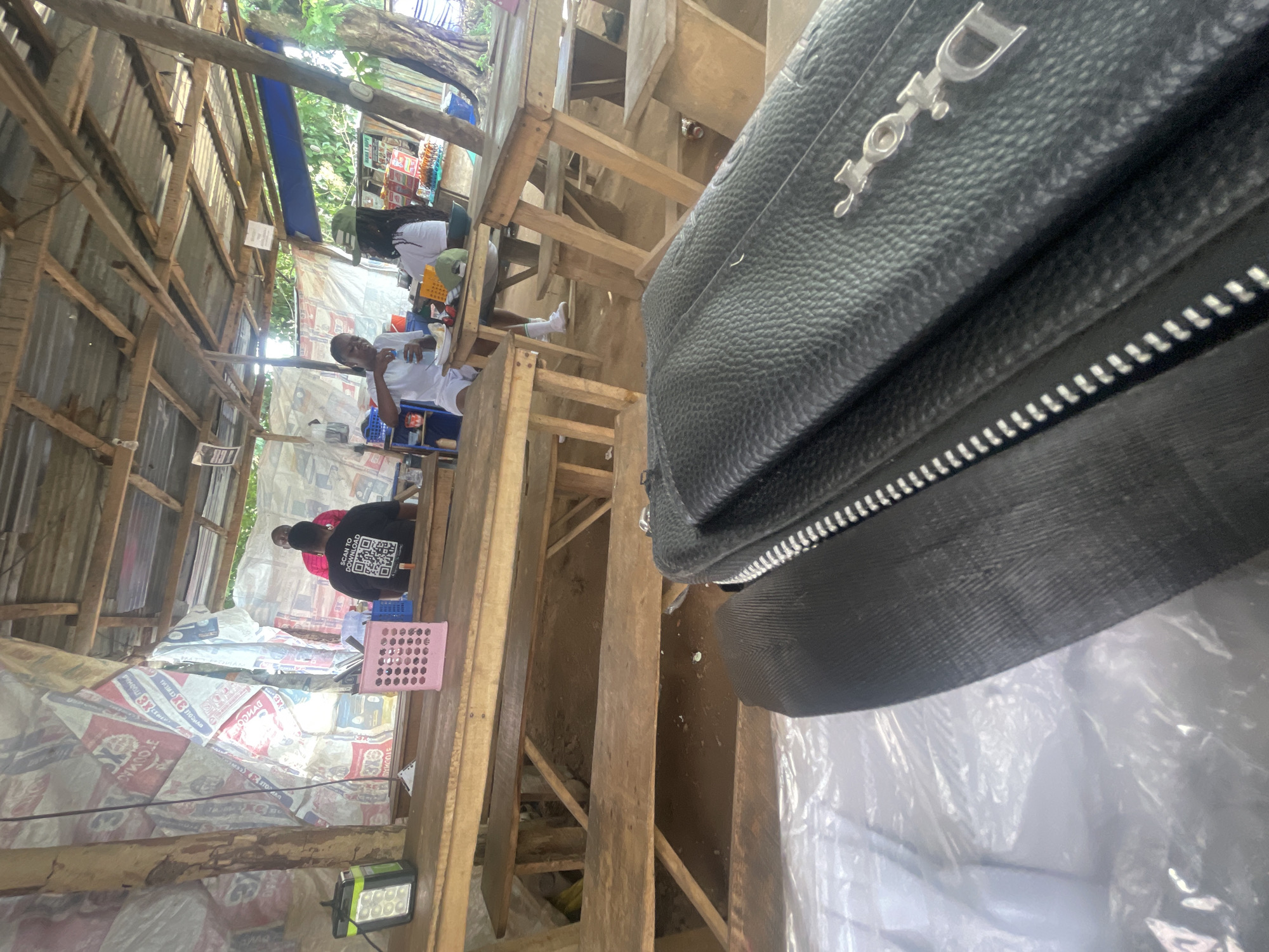 The image depicts an outdoor dining area, likely at a roadside establishment in Orun Ekiti, Nigeria. Several wooden tables and benches are arranged under a rustic, open-air shelter constructed with corrugated metal roofing and wooden beams. The walls of the shelter are draped with what appear to be tarpaulins or repurposed materials, some bearing text and graphics.
In the background, three individuals are visible. One person, wearing a black t-shirt with a QR code and the text "SCAN TO DOWNLOAD," is seated at a table. Another person, in a white shirt, is gesturing with their left hand while seated at a table. A third individual, also in a white shirt, is seated further back.
On the tables, various items are present, including cups, plates, a pink basket containing utensils, and bottles. To the left of the frame, a solar-powered lamp with multiple LED panels is placed on a table.
In the foreground, a black leather bag with a prominent "Dior" logo is placed on top of another object, partially obscuring the view of the dining area. The overall environment suggests a casual, possibly humid climate.