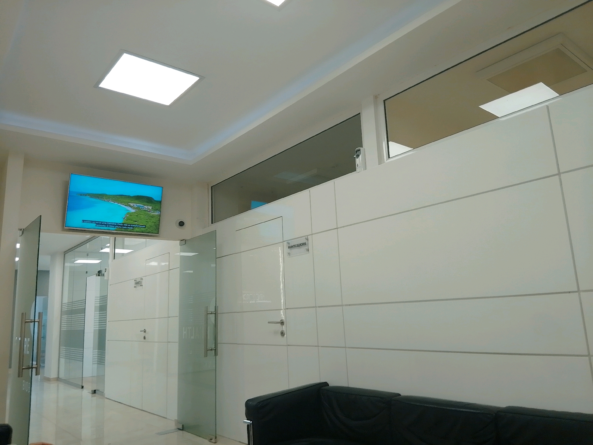 The image depicts the interior of a modern office or reception area. A large, flat-screen television displays a scenic image of a tropical coastline. The walls are tiled in white, with horizontal lines creating a grid pattern. Several glass doors with metal handles are visible, suggesting a series of offices or rooms. The ceiling is white and features a recessed, illuminated panel, and a narrow window runs along the upper portion of the wall, revealing a glimpse of what appears to be another room with similar lighting. A black leather sofa is partially visible in the foreground. The environment is clean, well-lit, and appears to be a professional setting. The text on the sign above the door reads "МАНТИЗАЦИОННА", which translates to "Sanitization". This suggests the location may be a medical or dental clinic. While the image itself does not provide specific information about its location in Sofia, Bulgaria, the presence of Bulgarian text on the signage indicates it is situated in a Bulgarian-speaking region.