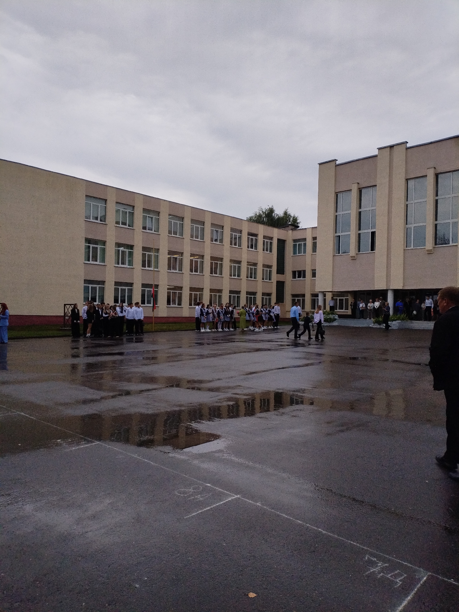 The image depicts a school assembly on a wet, overcast day in front of a large, multi-story school building. The building appears to be constructed of light-colored brick and concrete, with numerous windows. A group of students, predominantly in school uniforms, are gathered in formation in the courtyard. Some are standing in lines, while others are interacting in smaller groups. Two figures, a man in a dark suit and a person in a light blue outfit, are standing apart from the main assembly. The courtyard surface is asphalt and covered in puddles from recent rain, reflecting the building and the sky. Chalk markings are visible on the wet ground, including what appear to be numerical or directional indicators. The overall atmosphere is somber due to the weather and the formality of the gathering. The location is identified as a school in Bialynichy, Belarus.