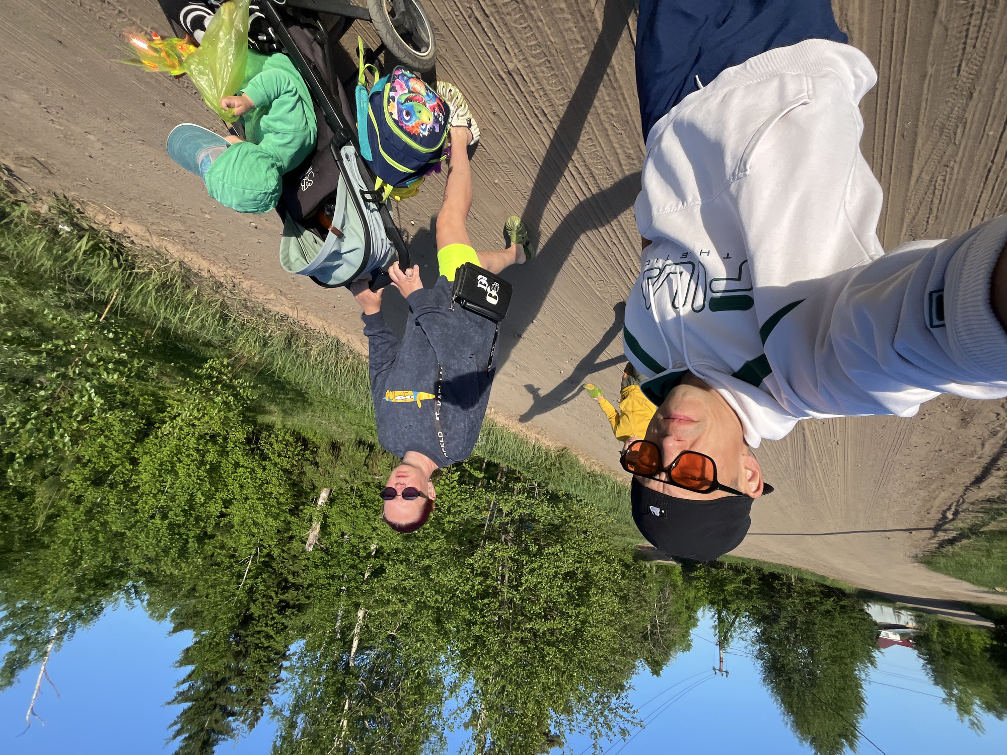 The image shows two adults and a child on a dirt road, likely in Chaikovsky, Russia. The adult on the right wears a white hoodie with green details, dark blue pants, a black baseball cap, and orange sunglasses. The adult on the left wears a blue-gray shirt, bright yellow shorts, and dark sunglasses. They are standing next to a stroller. A child in green clothing is in the stroller, along with a green plastic bag and a light blue bag. A blue and purple backpack is hanging on the stroller's handle. The surrounding environment includes trees, grass, and a blue sky. A building is visible in the distance. The photo appears to be a selfie taken by the adult on the right.
