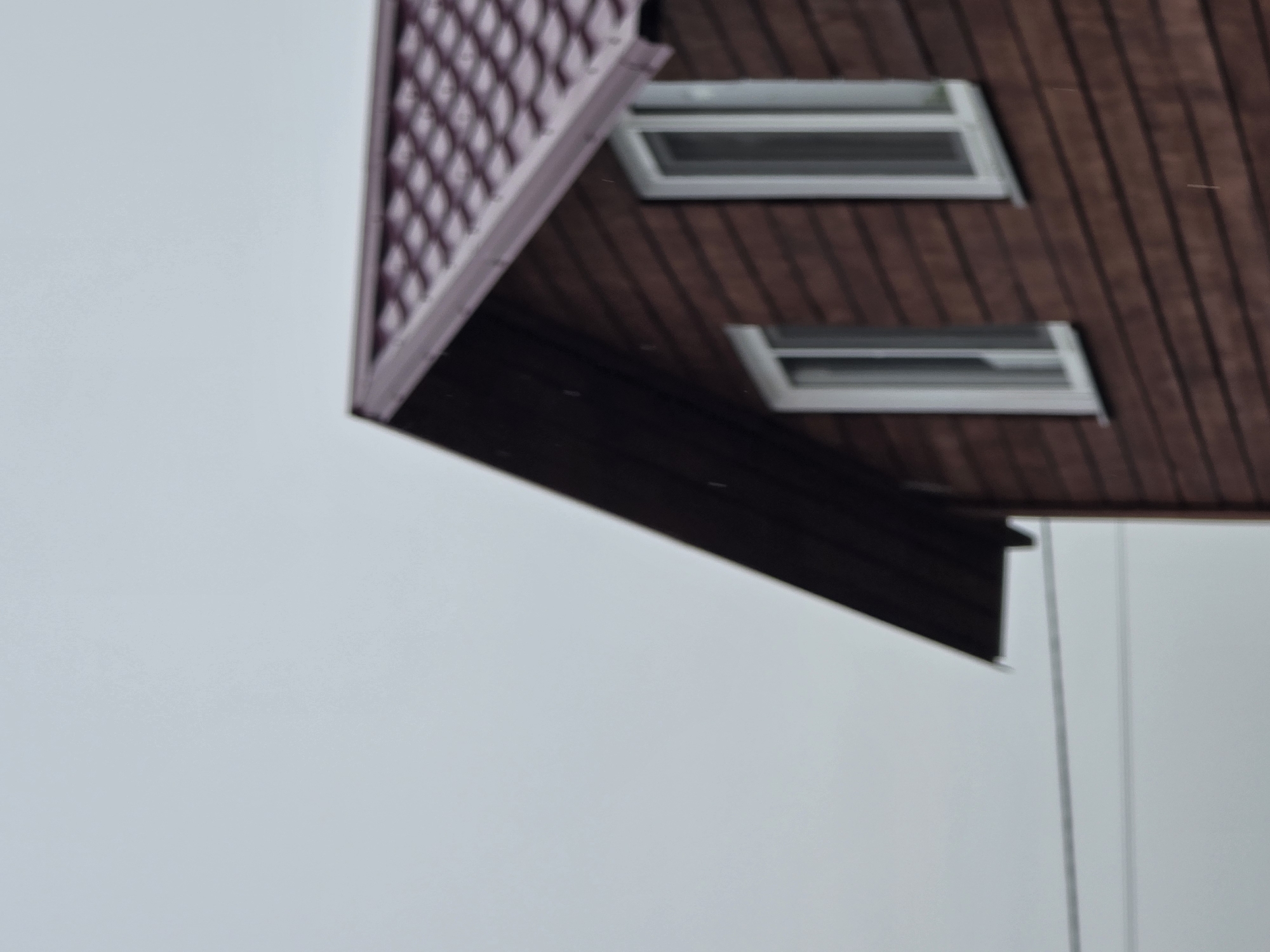 A blurred, medium-angle shot captures the upper section of a wooden house with a red, shingled roof under an overcast sky. The house has horizontal brown wood siding and two white-framed windows visible on the upper floor. A thin, dark wire stretches horizontally across the left side of the frame, suggesting its location in a populated area, possibly the city of Ермолаево, Russia. There are no people or animals visible.