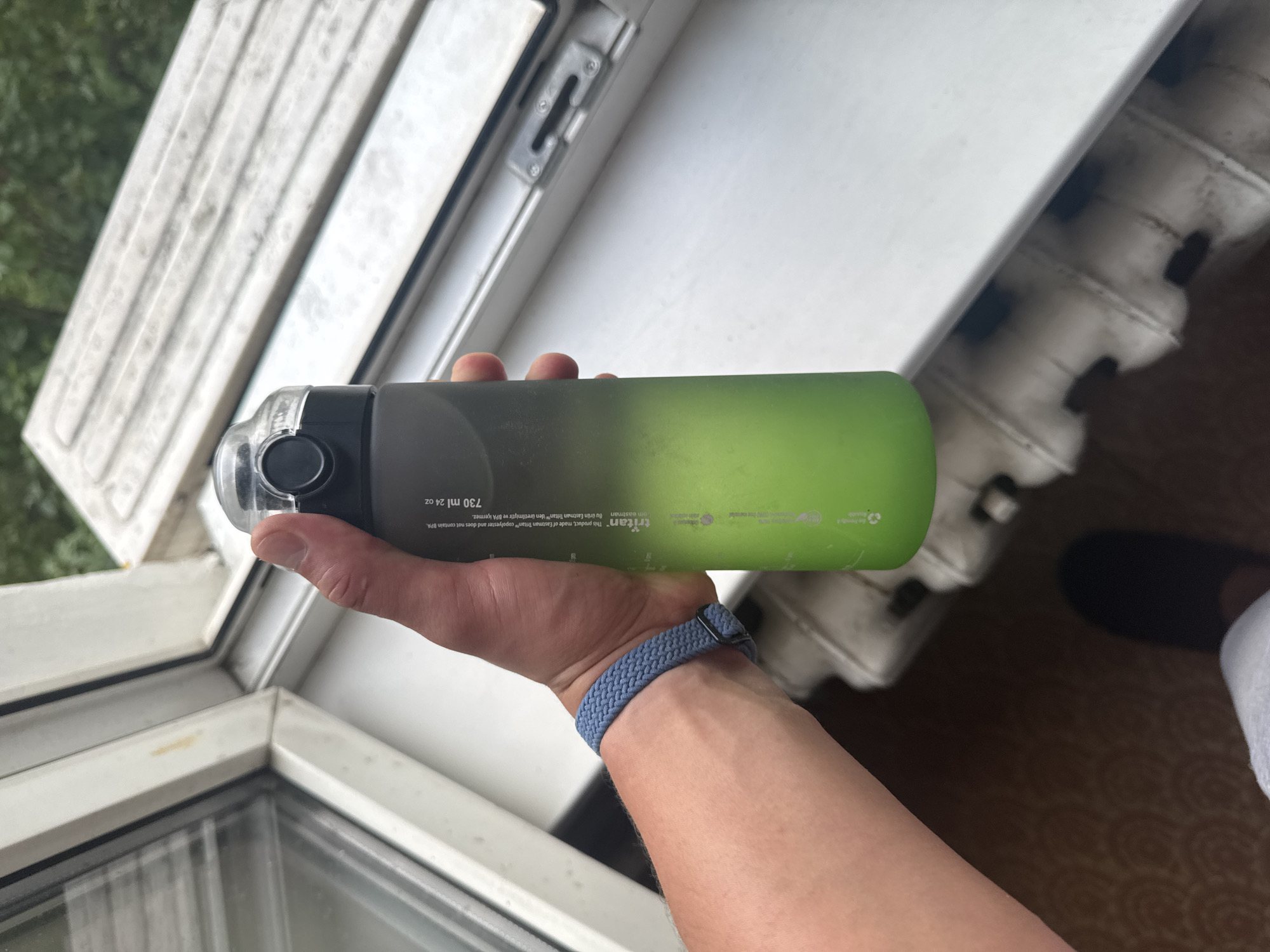 A person's hand holds a green and gray gradient water bottle with a black lid. The bottle has text printed on it, including "Tritan" and "750 ml 24 oz." A blue woven wristband is visible on the person's wrist. The background shows a white window frame and sill, a white radiator, and a glimpse of trees outside. The scene appears to be indoors, near a window.