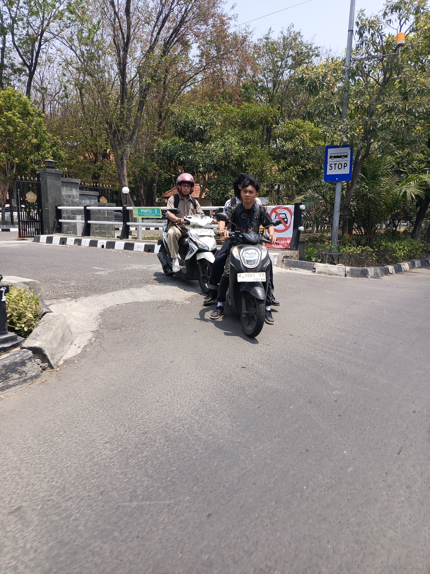 This image captures a street scene in Surabaya, Indonesia. Two motorcycles are visible, each carrying passengers. The motorcycle in the foreground is black, with a license plate that reads "W 3801 YA 02-30". It carries two young men. The man in front is wearing a black patterned shirt and a backpack. The man behind him is wearing a black t-shirt. The second motorcycle, to the left and further back, is white. It is carrying two people, one of whom is wearing a red helmet and a brown shirt, and a backpack. Both motorcycles are on a paved road.
In the background, there is a gate with intricate black metalwork, behind which are buildings with red roofs. A sign with the text "PINTU - 3" and some other Indonesian text is visible. There is also a traffic sign with the word "STOP" and a bus symbol, indicating a bus stop. Trees with green and some brown foliage line the street. The sky is clear and blue, suggesting a sunny day. The overall impression is a typical urban street scene in Indonesia.