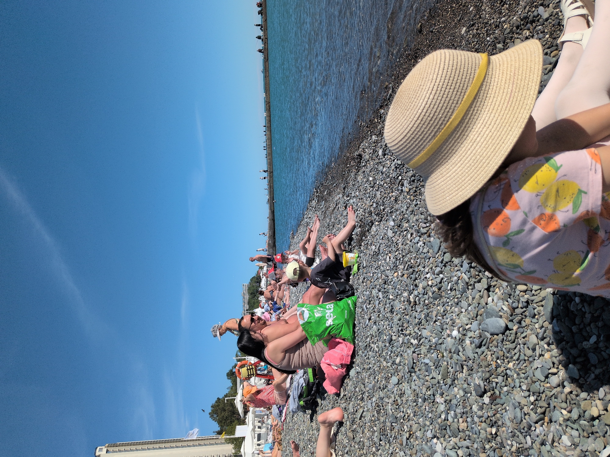 This image depicts a bustling pebble beach scene on a bright, sunny day, likely in Sochi, Russia. The perspective is slightly elevated, looking across the beach towards the sea and a distant pier.
In the immediate foreground, the back of a person, possibly a child, is visible. They are wearing a wide-brimmed straw hat with a yellow band and a light pink t-shirt adorned with cheerful lemon and orange patterns. Their legs, clad in white leggings and light sandals, extend towards the water's edge, resting on the smooth, grey and brown pebbles of the beach.
Further down the beach, numerous people are scattered across the pebbled expanse. Many are lying down, sunbathing, while others are seated or engaging in conversation. They are dressed in light summer attire or swimwear. A notable detail is a person with a dark top holding a bright green shopping bag, on which the brand name "Sela" is clearly visible. Several other individuals, including children, are present, suggesting a lively and family-friendly atmosphere.
The calm, blue sea stretches across the mid-ground of the image, meeting the pebbly shore. A long, grey pier or breakwater extends far into the water, with several small figures of people visible along its length. The sky above is a brilliant, cloudless blue, indicating clear weather. In the far distance on the left, white buildings, likely part of a coastal resort or city, are partially visible. The scene portrays a typical summer day of relaxation and leisure by the sea.