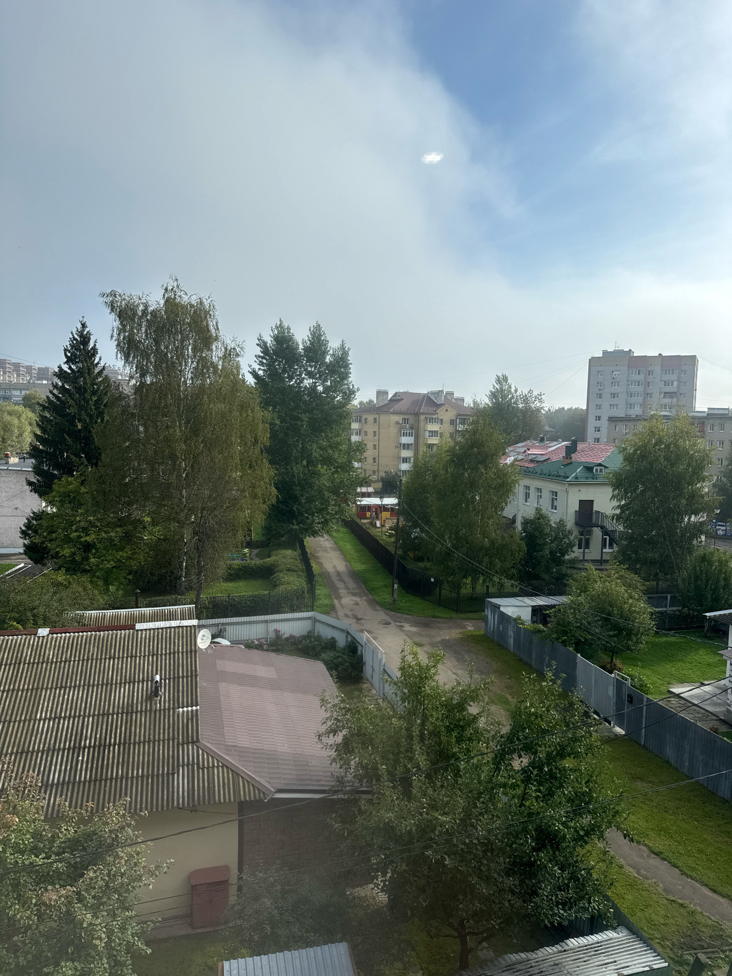 The image is an aerial view of a residential area in Rusanovo, Russia, likely taken from a multi-story building. The scene is dominated by buildings, trees, and a dirt road. In the foreground, there are two house roofs, one covered with corrugated metal sheets and the other with brown asphalt shingles. A satellite dish is visible on the latter. To the left, a building with a tan facade and a dark red outdoor storage unit is partially visible.
A dirt road winds through the middle ground, bordered by green grass and fences. Several trees, including tall evergreens and deciduous trees with green foliage, are scattered throughout the scene. In the background, apartment buildings of varying heights and architectural styles are visible, some with light-colored facades and red or green roofs. The sky is partly cloudy with patches of blue. The overall impression is one of a suburban or semi-urban environment with a mix of older and newer residential structures. There are no people or animals visible in the image.