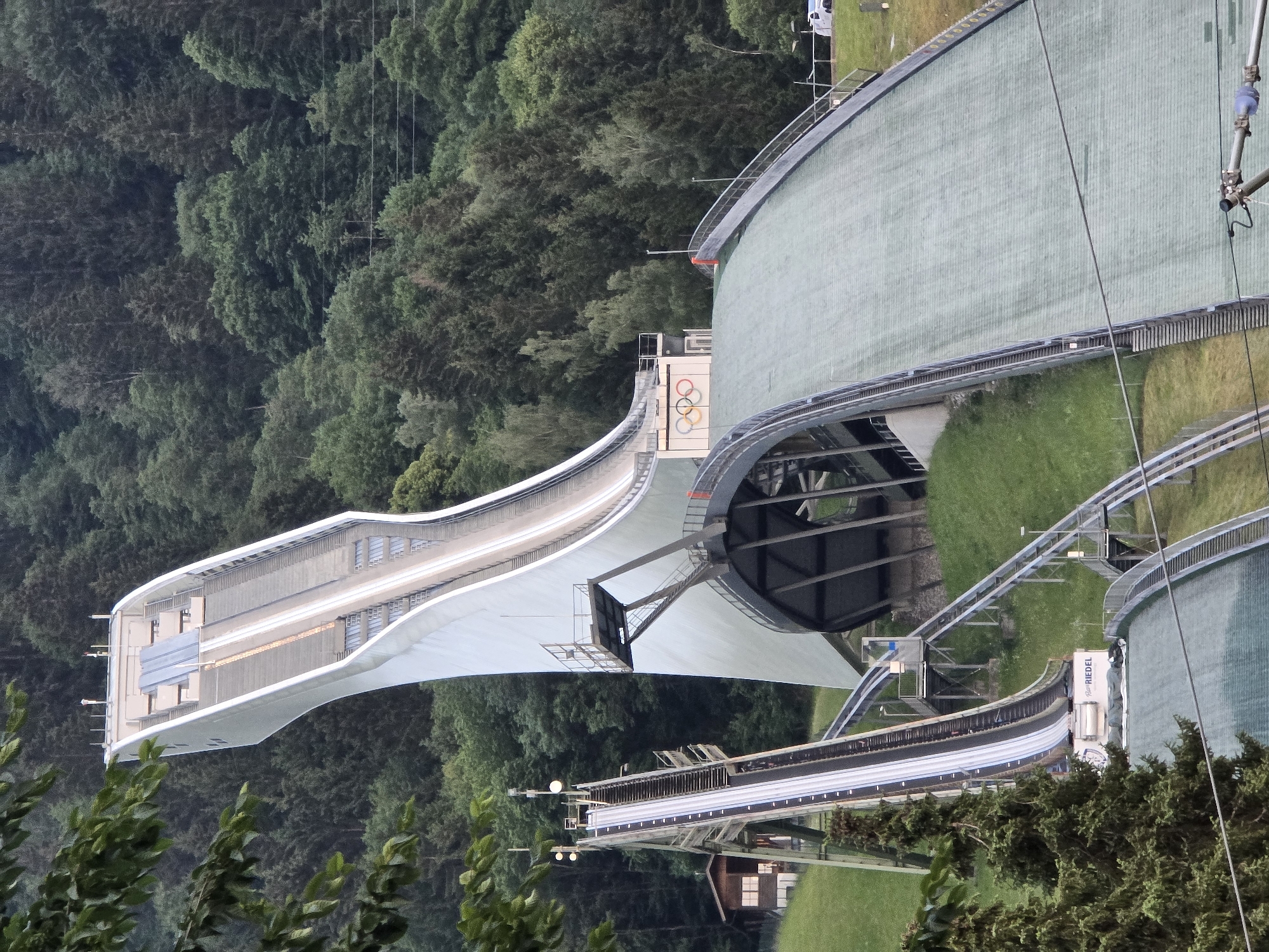 The image depicts the Olympic Ski Jumping Hill in Garmisch-Partenkirchen, Germany. The hill is a large, man-made structure designed for ski jumping competitions. It features a steep inrun track that athletes slide down, followed by a jump where they launch into the air. The landing hill is a sloped area covered with green turf, designed to safely absorb the impact of the jumpers.
In the foreground, the landing hill is visible, with railings along the sides. A section of the inrun track, made of metal and with visible tracks, ascends towards the top of the hill. A small wooden building is situated on the grassy slope to the left of the inrun.
The background is dominated by a dense, green forest that covers the surrounding hills. The sky is overcast, and the lighting suggests it is either early morning or late afternoon. The Olympic rings are visible near the top of the structure, indicating its historical significance. The overall scene conveys a sense of scale and the natural integration of the sporting facility into its alpine environment. There are no people visible in the image.
