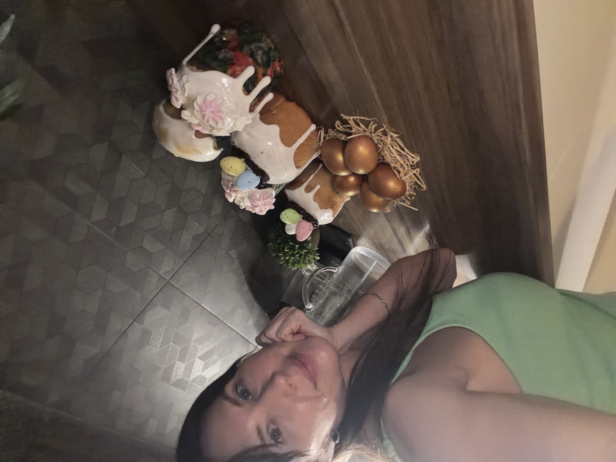 A woman is pictured in what appears to be a selfie, possibly taken indoors. She is fair-skinned with dark hair, wearing a light green top. A glass pitcher is visible near her arm. On a surface next to her, there are multiple decorated Easter cakes (kulich) with white icing and colored decorations. A nest-like arrangement with golden colored eggs is also present. The floor has a geometric pattern. The scene is located in Perm, Russia.