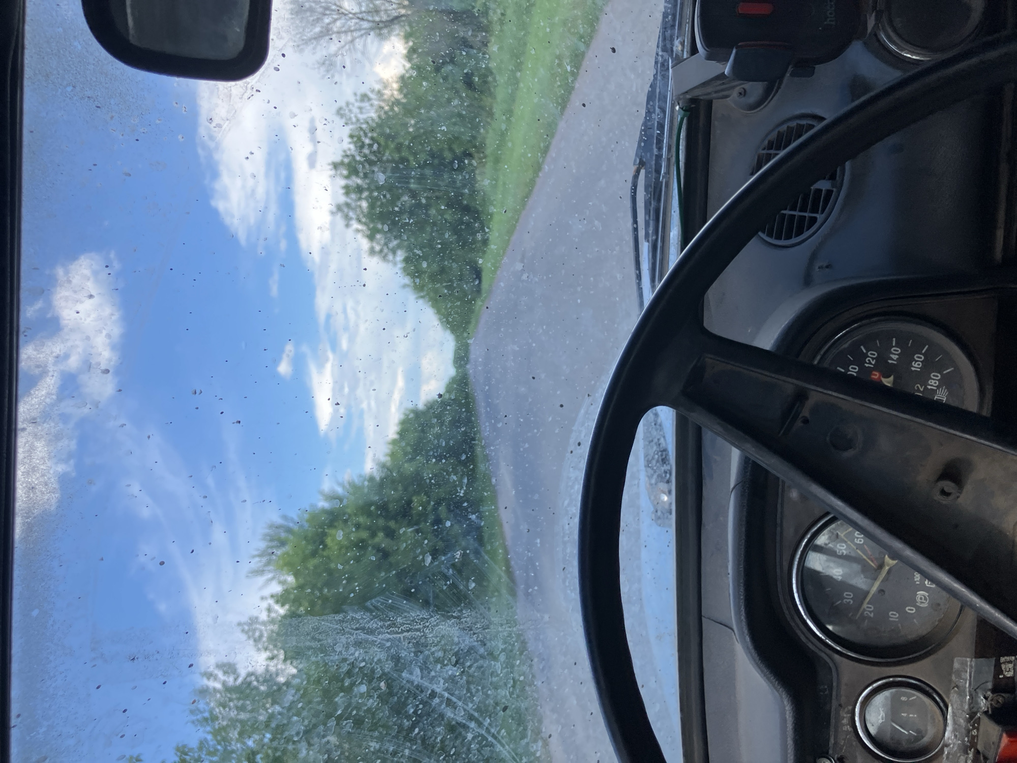 The image is a first-person perspective from inside a vehicle, looking forward through a dirty windshield. The vehicle appears to be a car, possibly an older model, with a dashboard visible in the foreground. The steering wheel, a circular black object, dominates the lower part of the frame. To the left of the steering wheel are two circular gauges, one with a needle pointing towards 30 and another smaller gauge to its left. To the right of the steering wheel, another gauge with a needle pointing between 140 and 160 is visible, along with a circular air vent.
The windshield is heavily splattered with what appears to be mud or water, obscuring the view to some extent. Outside the windshield, a paved road stretches into the distance, flanked by dense green trees on either side. The sky above is blue with scattered white clouds. The lighting suggests it is daytime, possibly late afternoon or early evening. The environment outside the vehicle appears rural, with no visible buildings or urban structures. The dirt on the windshield suggests the vehicle has been driven on a rough or muddy road, which is common in rural areas. The location is identified as Molodizhne, Ukraine.