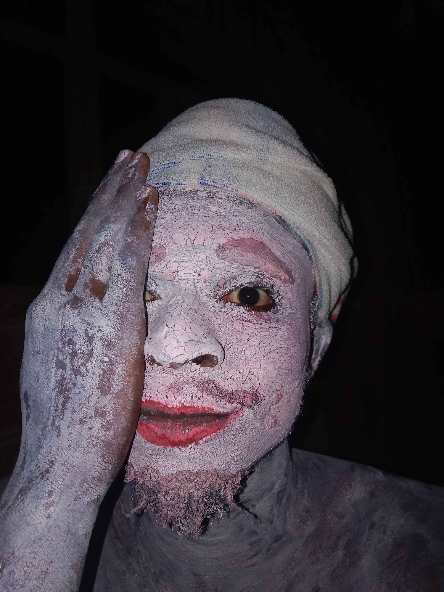 A person with a towel wrapped around their head is partially covered in a white, cracked substance and red lipstick, obscuring their features. Their hand, also covered in the white substance, is raised to their face. The individual's torso, visible beneath the neck, is coated in a similar substance. The background is dark and indistinct. There is no verifiable evidence to suggest this image was taken in Onitsha, Nigeria.