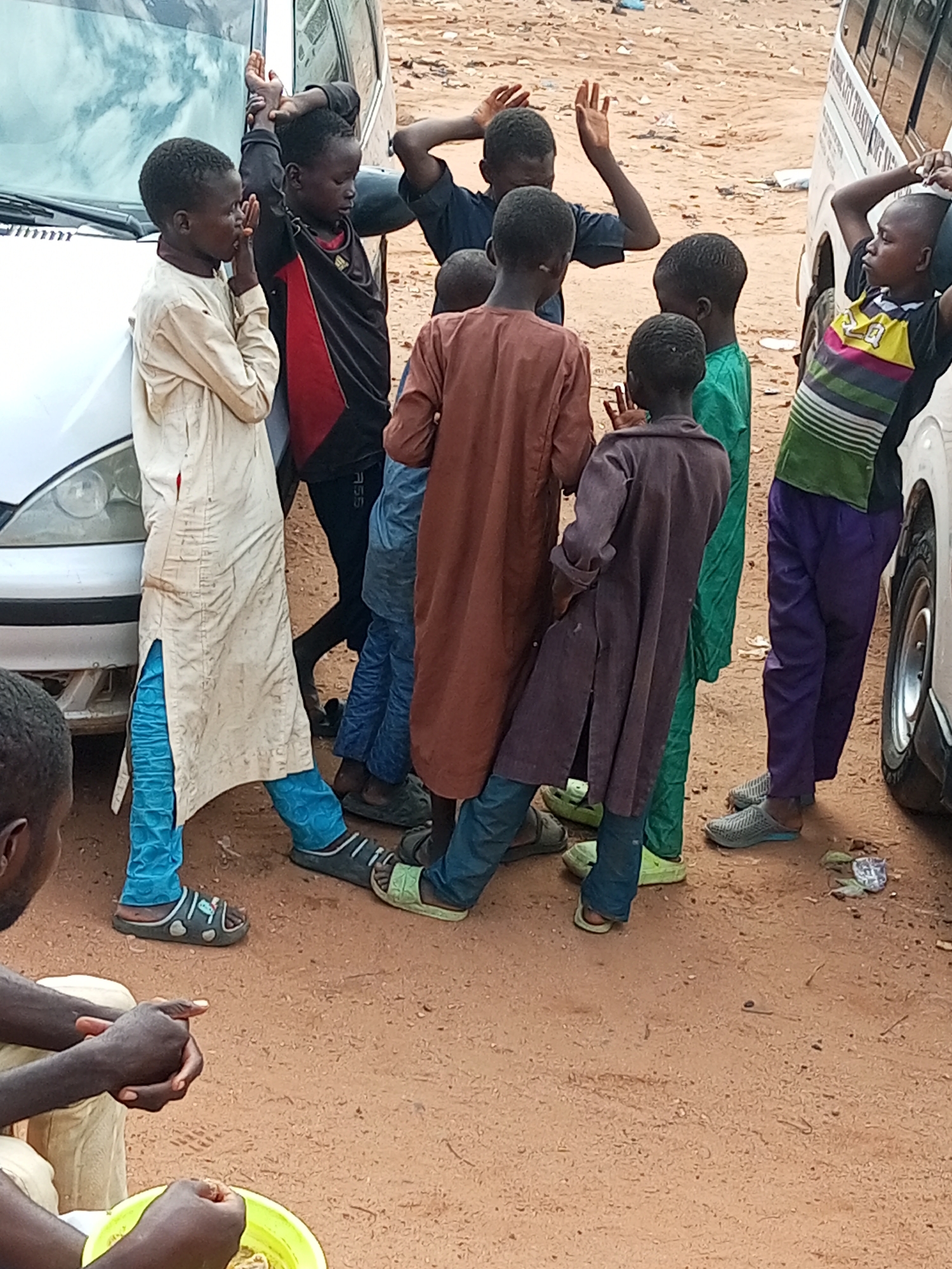 In a sandy, outdoor urban environment, a group of approximately ten boys, ranging in age from approximately six to sixteen years, are gathered around two vehicles. One vehicle, a light-colored sedan, is parked on the left side of the image, and a portion of a white van is visible on the right. The ground is covered in loose, brown sand with sparse debris. The boys are dressed in a variety of clothing, including traditional African attire such as long robes and trousers in shades of cream, purple, blue, and brown, as well as t-shirts and a black and red athletic top. Some of the boys are engaged in conversation, while others appear to be playing or interacting with each other. One boy, in a cream-colored robe and blue trousers, stands near the sedan with his hands clasped near his face. Another boy in a dark purple robe stands with his hands on his hips. The overall scene suggests a casual gathering in a semi-urban or informal settlement.