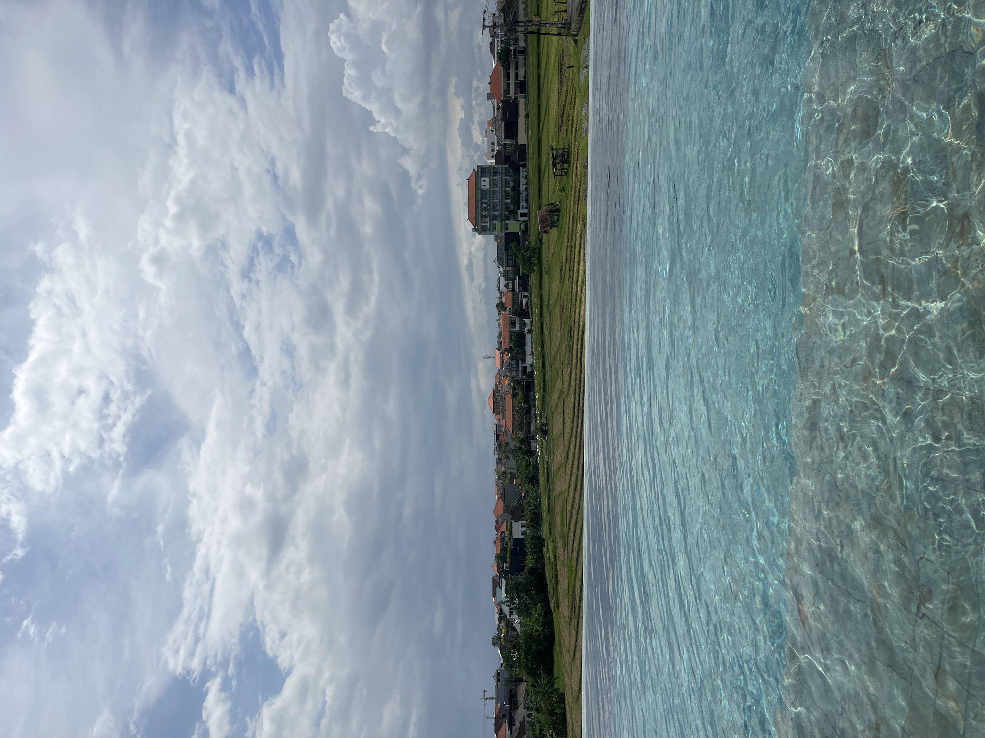 The image captures a scene in North Kuta, Indonesia, featuring a partially visible infinity pool in the foreground, with a view extending to a residential area and the sky. The pool's water appears light blue and rippled. Adjacent to the pool is a strip of land displaying a field or grassy area. Beyond this, there's a row of buildings of varying heights, with a mix of residential and commercial structures. The sky above is filled with white and gray clouds, suggesting a partly cloudy day. There are no people visible in the shot. The overall impression is a blend of leisure and the local architecture against a cloudy sky.
