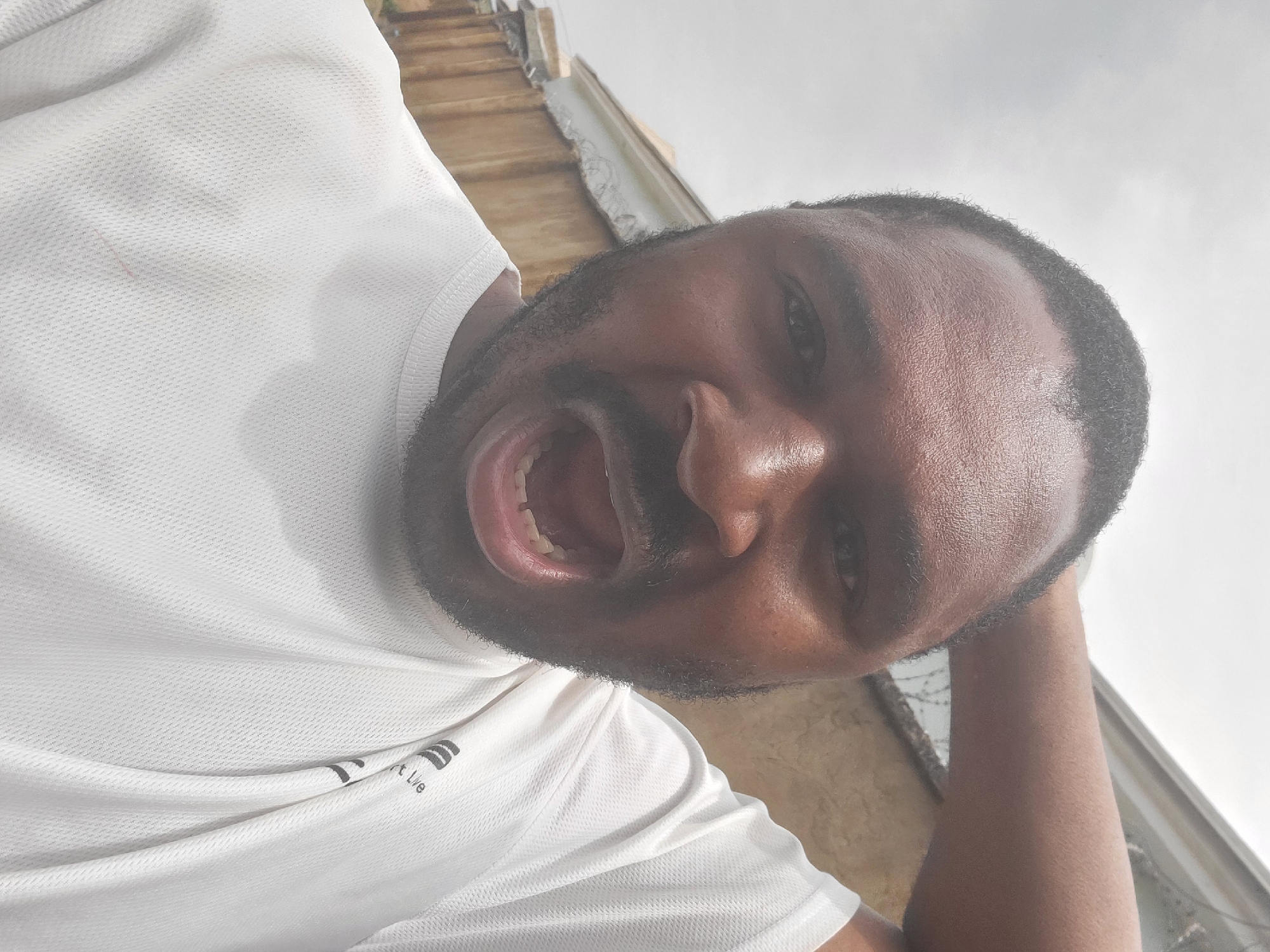 A close-up, rotated image shows a dark-skinned Black man with a beard and short hair, wearing a white t-shirt with a logo that reads "Smart Life". The man has his mouth open wide. His right arm is raised and resting on the back of his head. In the blurred background, there is an outdoor stairwell on a building with a razor wire visible. The location is Suleja, Nigeria.