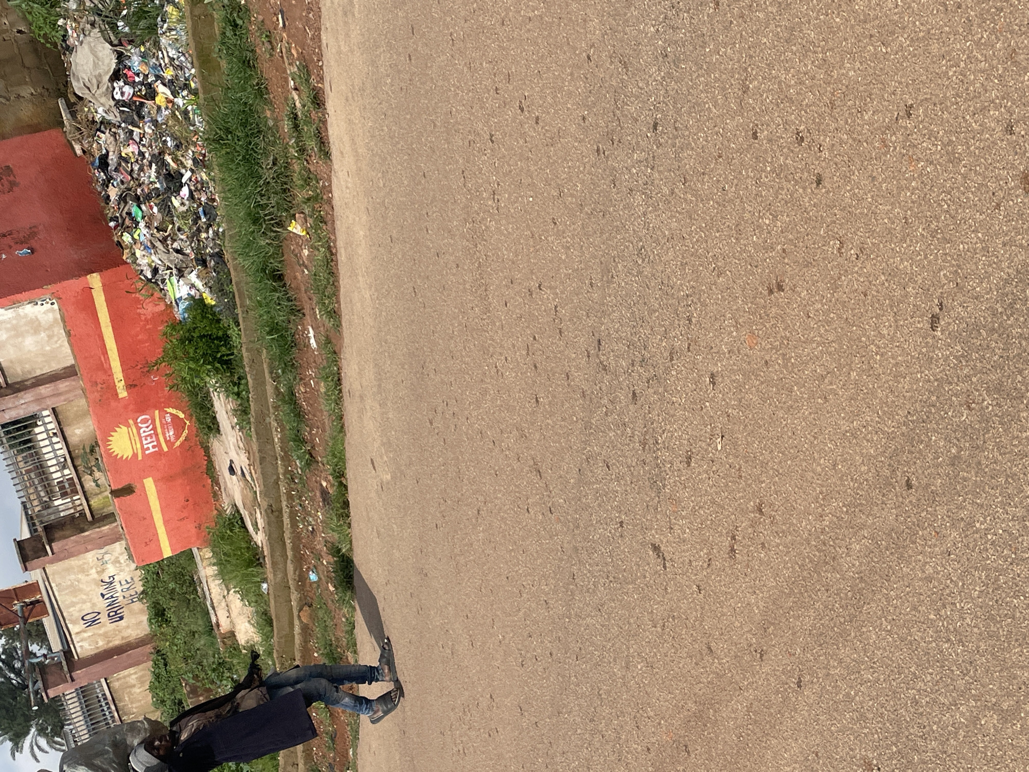 A person is walking on a paved road in Jos, Nigeria. The person is wearing a dark coat over a light-colored shirt and blue jeans, with dark flip-flops on their feet. They are carrying a large, light-colored object over their left shoulder. Behind the person, a red structure with a yellow stripe is visible, with the word "HERO" and a logo printed on it. Next to the red structure, a large pile of trash is accumulated. In the background, there is a building with a barred fence and some greenery. The ground is paved, and there is a curb with some grass and dirt on the side.
