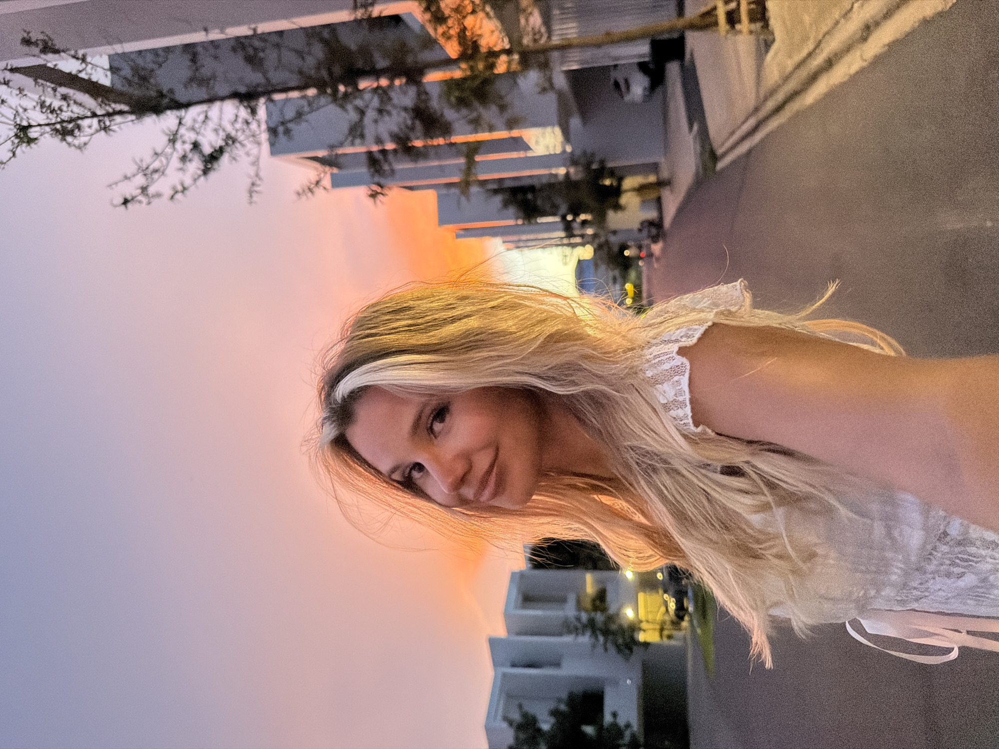 This is a selfie of a young, blonde woman with long, wavy hair, wearing a white lace top. She is smiling and looking at the camera. The background shows a modern, white building with several stories and some greenery. The sky is a gradient of purple and orange, suggesting it is either sunrise or sunset. The scene appears to be an outdoor setting, possibly a street or walkway, with asphalt visible in the foreground. The lighting indicates it's either dawn or dusk. There is no visible text in the image.