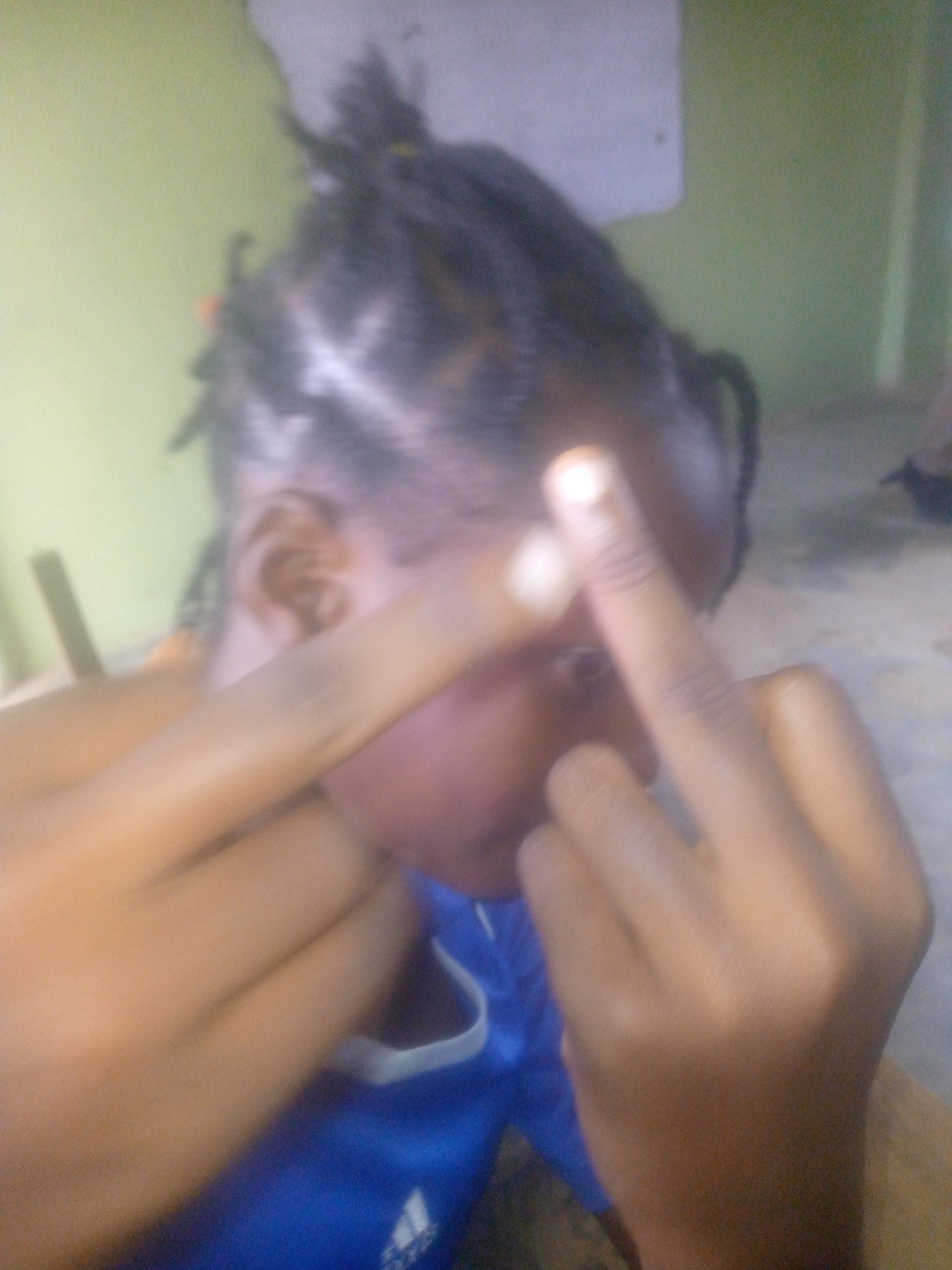 A person with dark skin and braided hair is obscuring their face with their hands, giving the middle finger. The person is wearing a blue Adidas shirt. The background is a light green wall, possibly indoors. The location is not explicitly discernible as Ikorodu, Nigeria from the image.