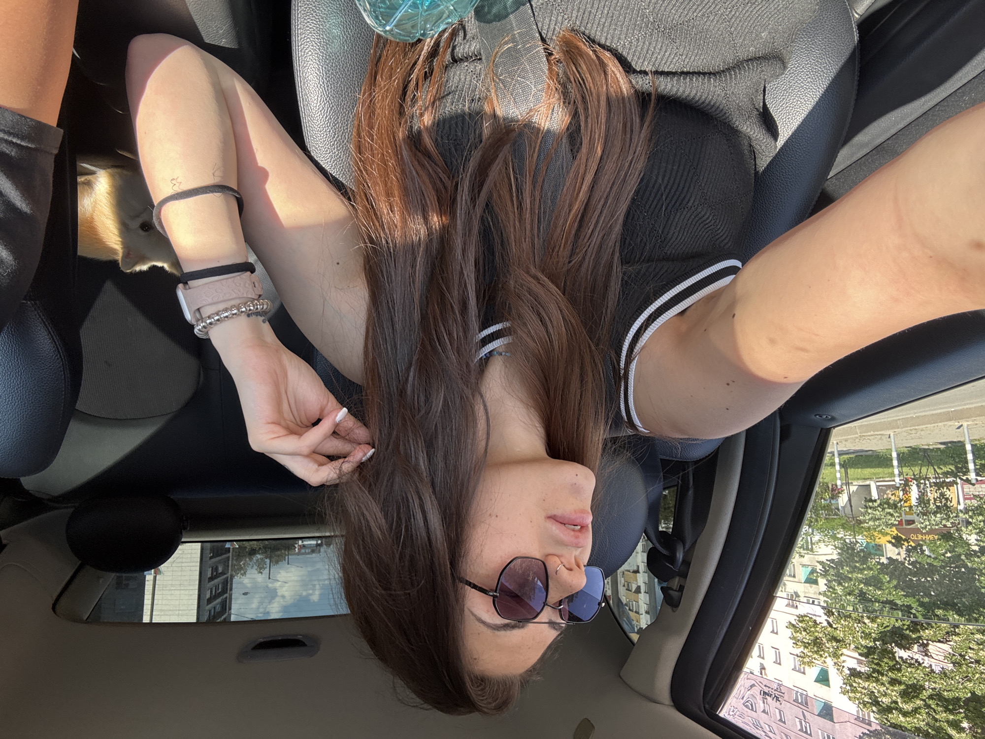 A young woman with long brown hair and sunglasses sits in the passenger seat of a car. She is looking to her right, away from the camera, and has her hand raised to her hair. The interior of the car is visible, including the black leather seats. A small, fluffy, light-colored dog is visible in the back seat behind her. Outside the car window, apartment buildings and trees can be seen, suggesting an urban environment. The scene appears to be taking place on a sunny day.