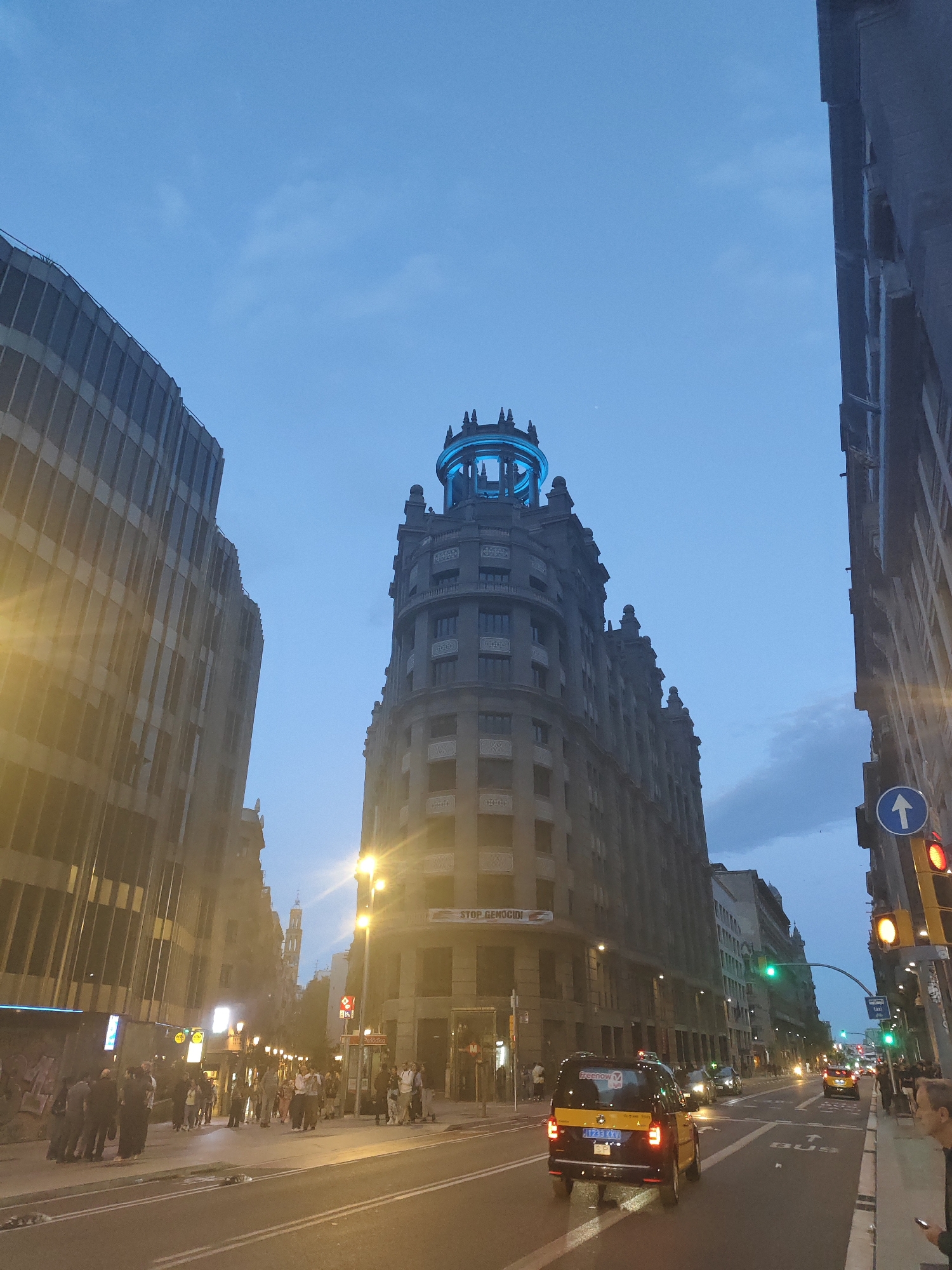 Here's a detailed description of the image:
**Setting:** The image is taken on a street in Barcelona, Spain. The architecture, street layout, and the "STOP GENOCIDE" banner (if it is present) suggest this is a demonstration taking place on a major thoroughfare.
**Objects and Infrastructure:**
* Tall buildings flank the street on either side. The building with the blue-lit structure at the top is distinctive.
* A traffic light displays a red signal.
* Street lights illuminate the scene.
* A taxi cab is driving in the street.
* A "BUS" marking is visible on the road.
**People:**
* Groups of people are gathered on the sidewalks. Some are on the left sidewalk. They appear to be a mix of pedestrians and possibly participants in a gathering.
* One person is seen to the right, standing on the sidewalk.
**Action:**
* Vehicles are traveling along the street.
* People are present on the sidewalks, likely walking or observing.
**Environment:**
* The sky indicates it is either dusk or early evening.
* The light conditions suggest it is artificial lighting that is illuminating the street.
**Location Clues:**
* The architectural style of the buildings points to a European city, specifically Barcelona.
* The presence of taxi cabs reinforces a city setting.
* The banner reading "STOP GENOCIDE" may indicate a protest or demonstration, giving further clues to the environment.