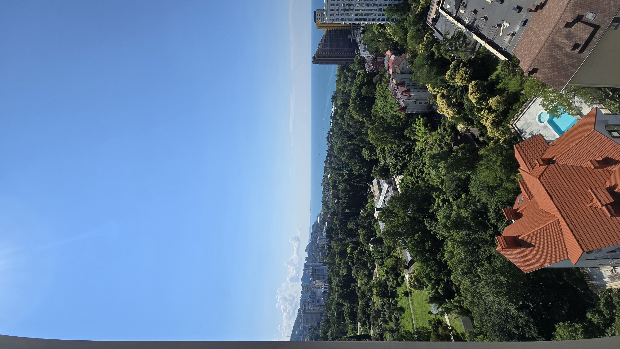 A high-angle, medium-shot captures a panoramic view of a coastal city, likely Sochi, Russia, under a clear blue sky. The foreground is dominated by the terracotta-tiled roof of a large residential building. To the right, the flat, dark rooftop of another building with visible HVAC units and infrastructure is partially visible. Beyond these structures, a lush, dense green forest stretches across the landscape. Scattered within the forest are various buildings, including smaller residential homes with red-tiled roofs and a distinct castle-like structure with turrets. Further in the distance, a cluster of taller, modern buildings and high-rises form a city skyline. The scene culminates with the expanse of the tranquil, blue sea meeting the horizon, with a few white clouds dotting the sky and small boats visible on the water. The environment appears to be a developed urban area integrated with significant natural greenery and proximity to the sea.