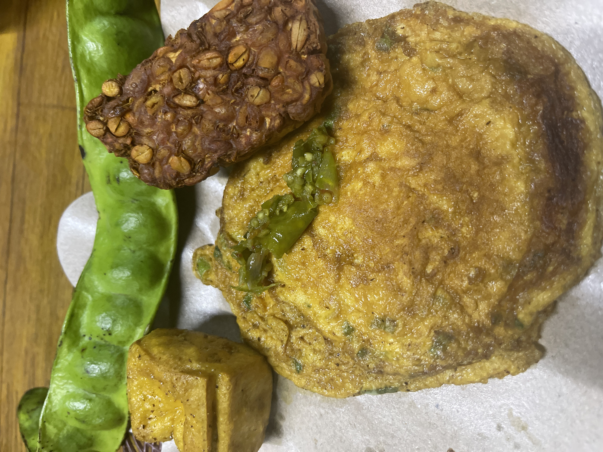 The image displays a close-up, overhead view of a meal laid out on a piece of textured paper, likely a serving mat, placed on a wooden surface. The meal consists of several distinct food items. In the foreground, a large, golden-brown omelet occupies a significant portion of the frame. It appears to be seasoned with finely chopped green ingredients, possibly scallions or herbs, and topped with a few pieces of sliced green chili. To the left of the omelet, a golden-brown, cube-shaped piece of fried tofu is visible. To the right of the omelet and slightly behind it is a piece of fried tempeh, a fermented soybean product, characterized by its dark brown, textured surface with visible bean fragments. Positioned in the background, arching across the top of the frame, is a pod of green beans, likely petai (stink beans), identifiable by their plump, segmented appearance. The overall lighting suggests an indoor setting, possibly a home kitchen or a casual eatery in Garut, Indonesia, a region known for its diverse culinary traditions.