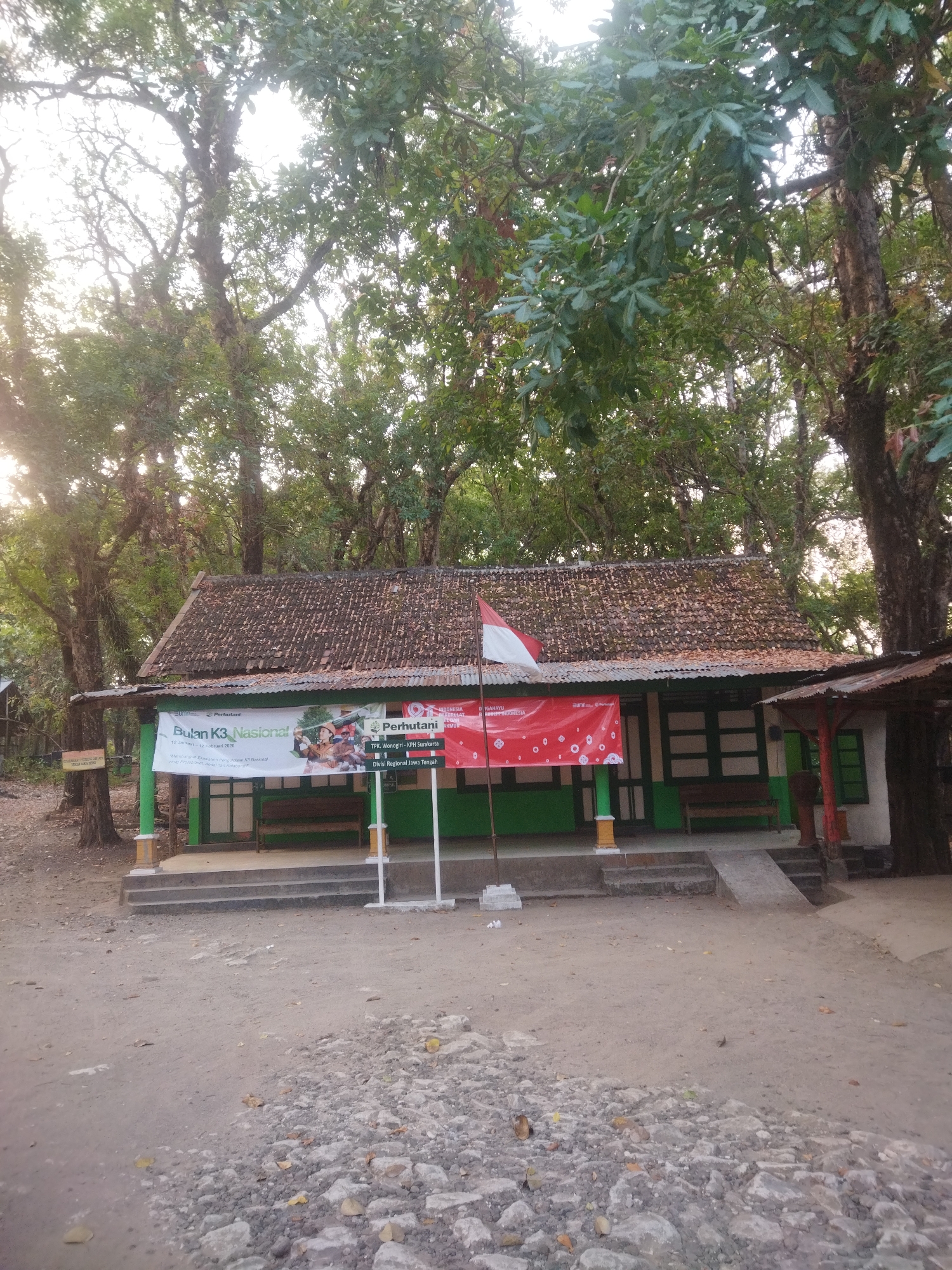 The image depicts an outdoor scene in Wonogiri, Indonesia, featuring a small building with a tiled roof and green trim. In front of the building are two banners. The banner on the left is white with green text and imagery, while the banner on the right is red with white circular patterns and text. A flag pole with an Indonesian flag is positioned between the banners. The building appears to be part of a local establishment or administrative office, with a bench visible inside. The surrounding environment consists of sandy ground, rocks, and mature trees. The overall impression is one of a rural or semi-rural setting, likely a public space.