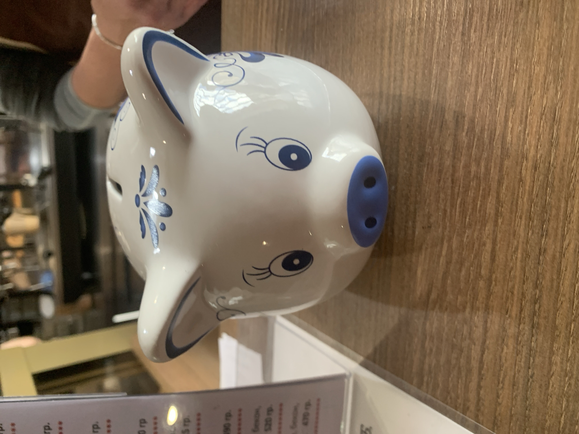 A white ceramic piggy bank with blue floral accents and blue eyes and nose sits on a wooden table. A menu or price list is partially visible to the left of the piggy bank. In the background, out of focus, a person's hands and arms are visible, suggesting they are seated at the table. The environment appears to be an indoor setting, possibly a cafe or restaurant. The time of day cannot be determined. The location is identified as Pastavy, Belarus.