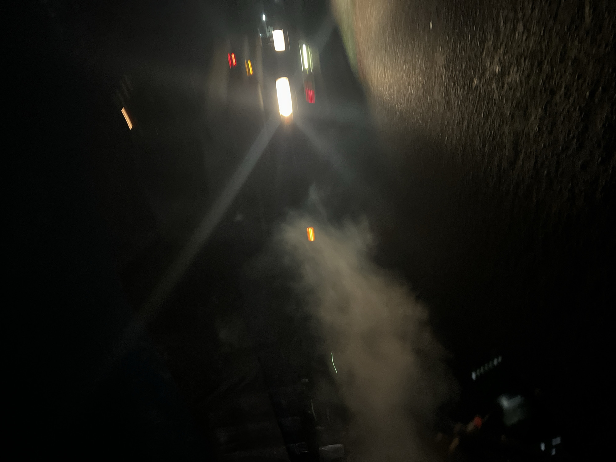 The image depicts a night scene on a road in Kubwa, Nigeria. Several vehicles with illuminated headlights and tail lights are visible in the distance, partially obscured by smoke or fog. The road surface is dark and appears wet. The right side of the image shows a textured surface, possibly a wall or embankment. The overall lighting is poor, with limited visibility due to the darkness and atmospheric conditions.