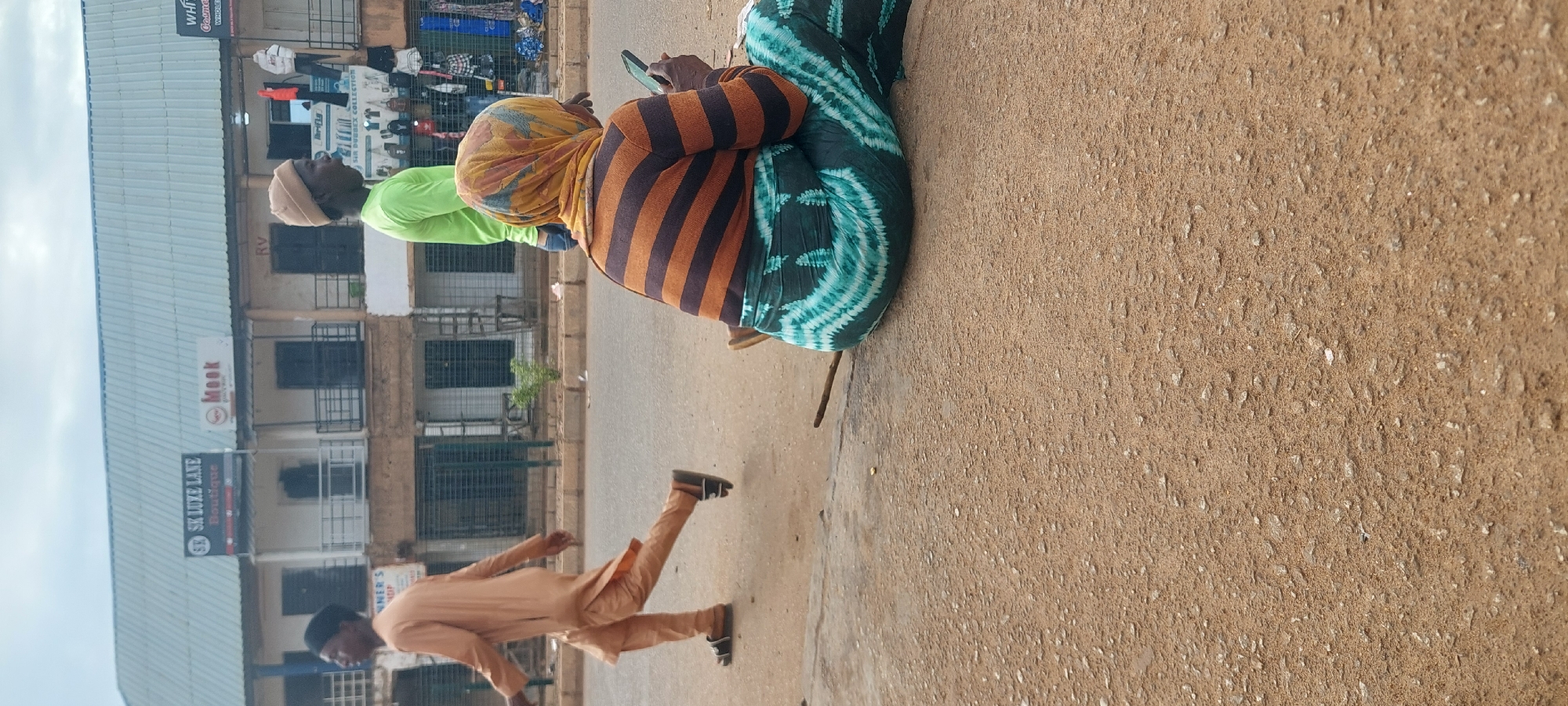 This image depicts an outdoor street scene in the city of Unknown, Unknown. In the foreground, a rough dirt and gravel surface occupies the lower half of the frame. Towards the right, an individual is seated directly on this ground, viewed from behind. This person, possibly an adult female, wears a horizontally striped long-sleeved top in dark brown and orange, a patterned long skirt in dark green and teal, and a multi-colored headscarf featuring yellow, orange, and brown tones. They are holding a dark-colored smartphone and looking down at it. A thin wooden stick lies on the ground near the seated individual.
In the middle ground, a paved road stretches horizontally across the frame. Two male figures are visible on or near the road. On the left, a man in a light brown tunic and matching trousers, wearing dark sandals, walks from left to right. Further right, a second man wears a bright lime green long-sleeved shirt and a light tan beanie, facing towards the left.
The background features a row of two-story commercial buildings with light grey corrugated metal roofs. Visible signage includes "SK LUXE LANE Boutique" and "Meek" on the left, and partially obscured signs for "RV," "WHITE Cosmetics," and "SIR DUBBEX COLLECTION" further right. Various items of clothing and merchandise are displayed on racks outside some of the shops. The sky is overcast.
