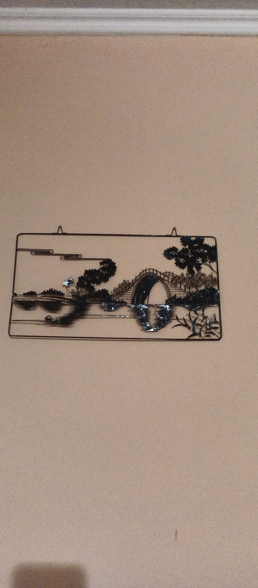 This is a close-up, eye-level, medium shot of a metal wall art piece hanging on a tan wall. The art depicts a traditional East Asian landscape with a bridge arching over water. Trees and foliage are rendered in a silhouetted style, with reflective highlights on the water suggesting movement. The wall art is framed by a thin black border and suspended by small hooks at the top. Above the art, a white decorative molding runs along the top edge of the wall. The lighting is soft and casts a slight shadow. There is no indication of the location within the city of Berehove, Ukraine.