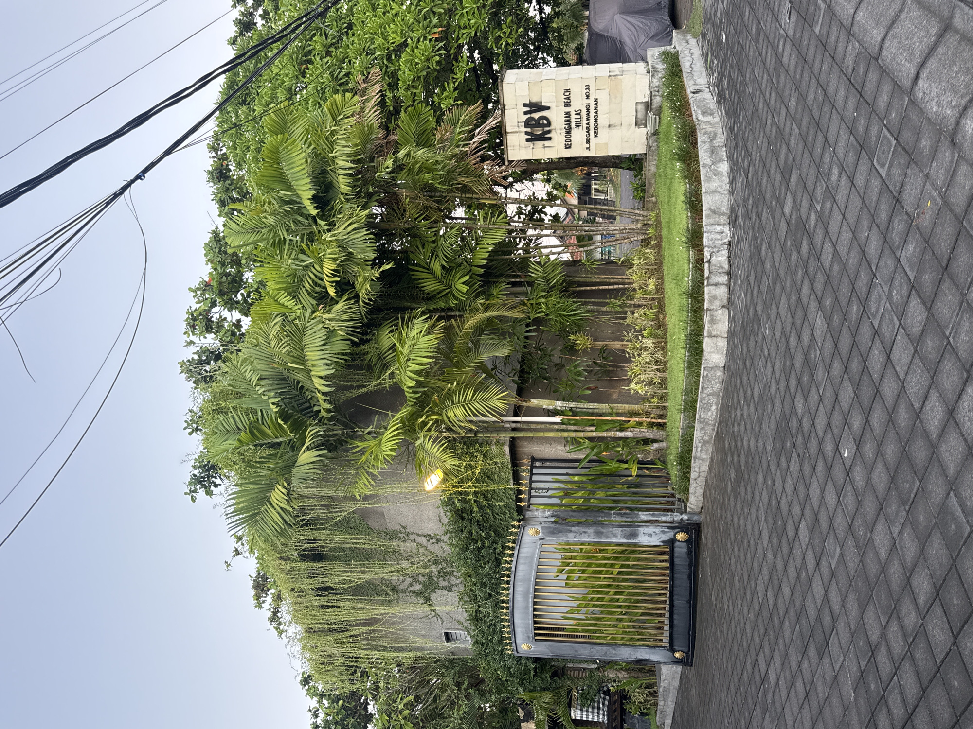The image shows the entrance to Kedonganan Beach Villas (KBV) in Kuta, Indonesia. A stone sign displays the name of the villas and the address Jl. Segara Wangi No.33, Kedonganan. Lush green foliage and trees frame the entrance. A black metal gate with gold-colored accents stands slightly ajar. The building behind the gate is partially covered in climbing plants. Overhead power lines are visible against the sky. The foreground is a paved area with interlocking gray blocks. A street light is visible in front of the gate.