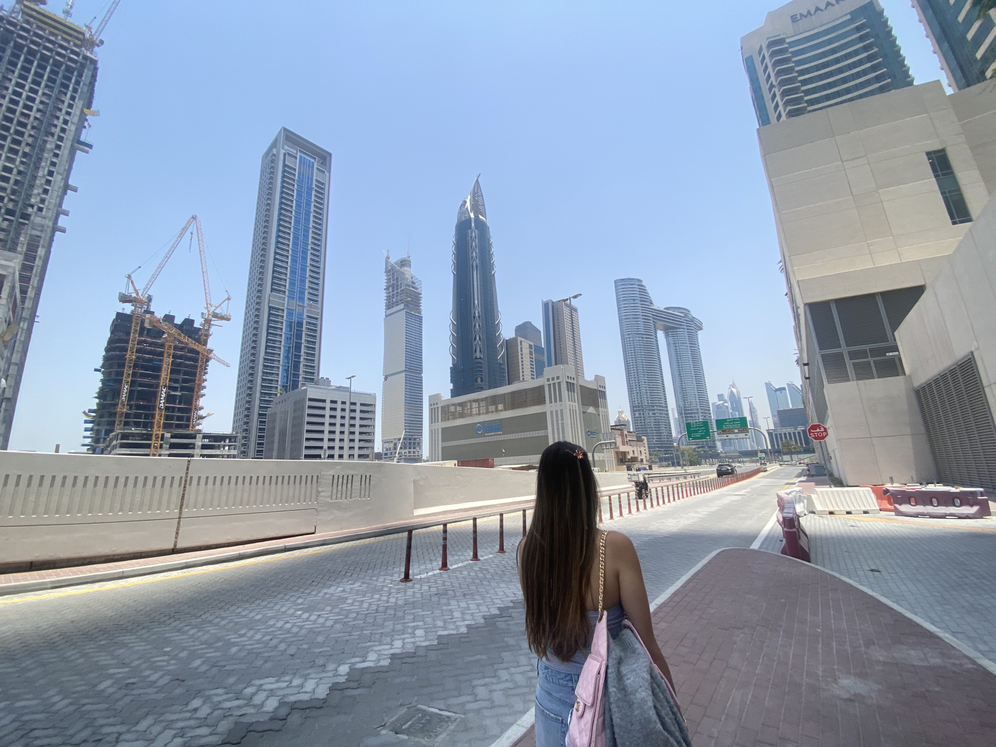 A woman is standing on a wide, paved street lined with metal barriers, looking towards a cluster of modern skyscrapers in Dubai, United Arab Emirates. Some of the notable buildings visible include a tower under construction with multiple yellow cranes, the blue-windowed Almas Tower, the curved twin towers of Dubai Canal, and a building with "EMAAR" emblazoned on its upper section. A "STOP" sign is visible on a building adjacent to the road. Traffic is sparse, with a few vehicles visible in the distance. The woman is wearing a tank top and jeans, with a pink bag and a gray jacket slung over her shoulder.