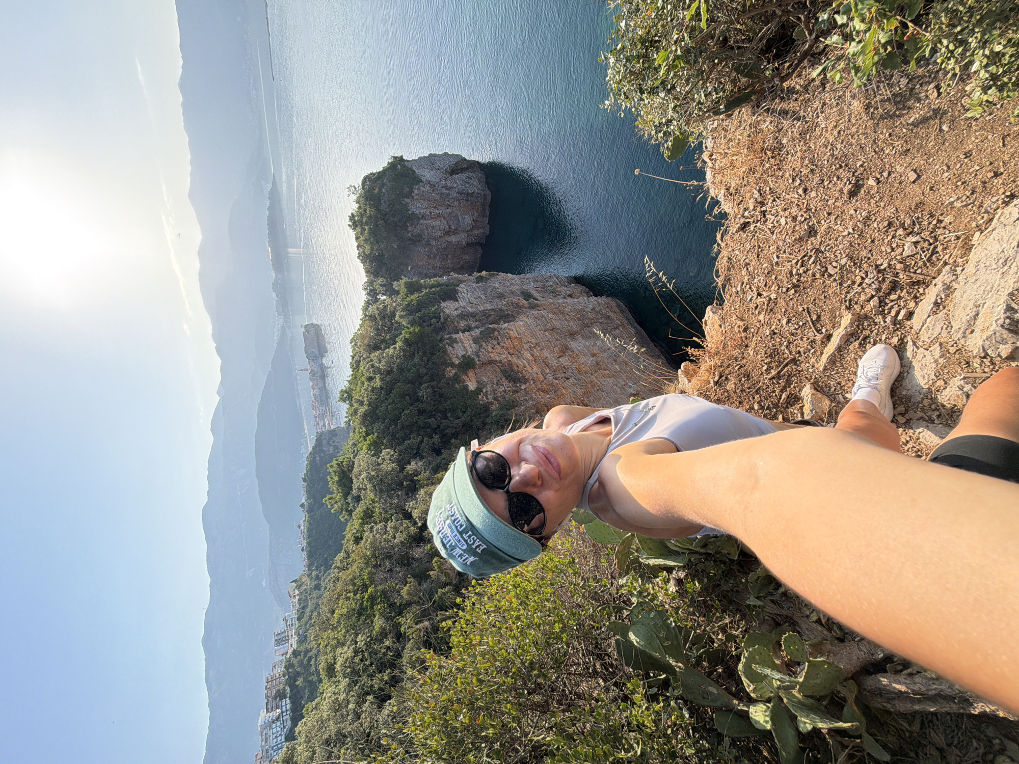 A woman takes a selfie while standing on a cliff overlooking the Adriatic Sea. She is wearing a green baseball cap, sunglasses, a light gray tank top, and white sneakers. Behind her, the rugged coastline of Budva, Montenegro, is visible, with buildings perched on the hillsides and a clear blue sky above. Two rocky islets rise from the water in the middle ground. The cliff edge where she stands is covered in dry grass and small stones.