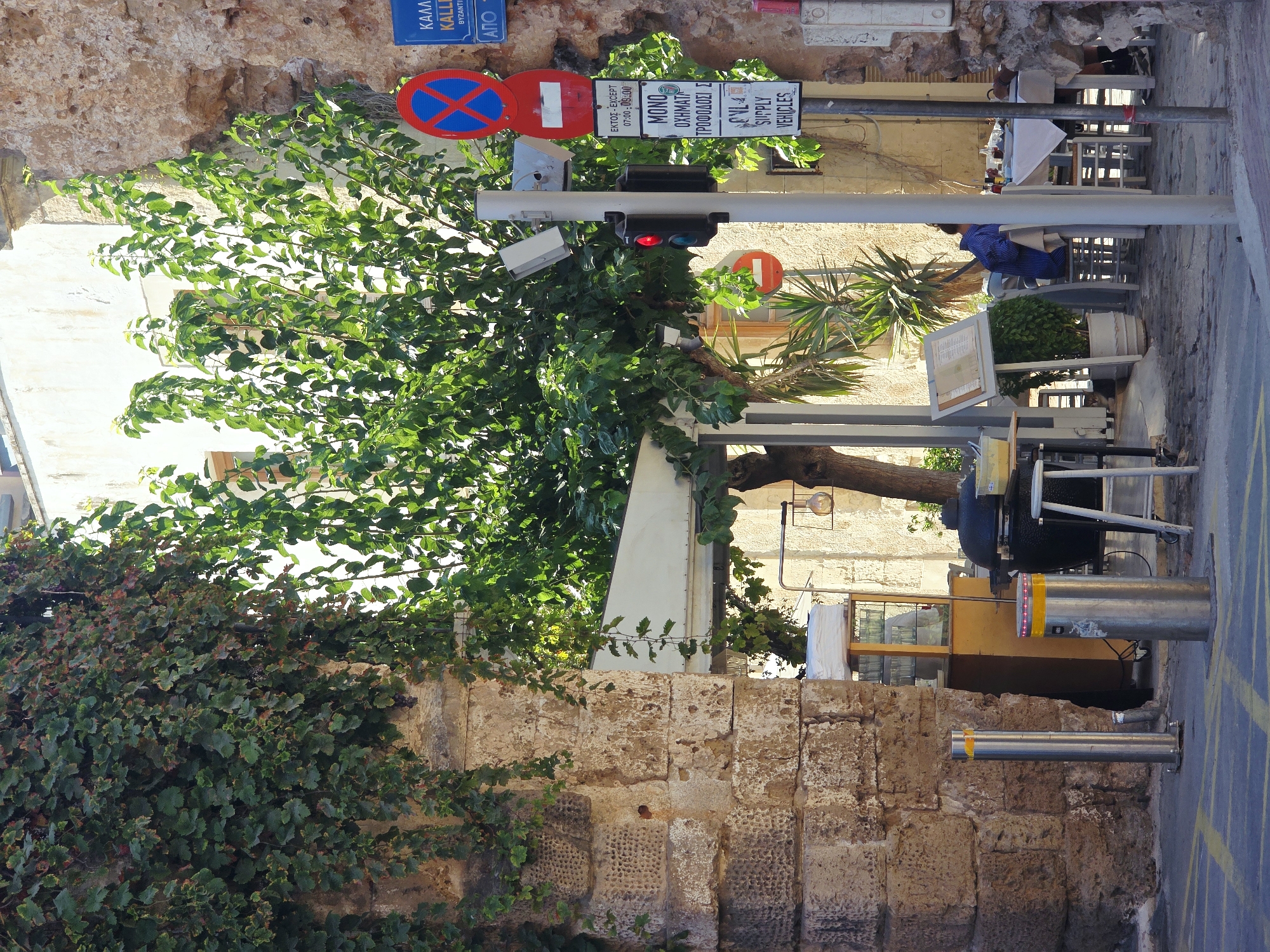 The image captures a street scene in Chania, Greece, focusing on an area with outdoor dining and historical architecture. In the foreground, to the left, is a weathered stone archway covered in dense green ivy. To the right of the arch, a metal bollard with yellow reflective tape stands on the paved street. Further into the scene, a traffic light with a red signal is visible, mounted on a pole along with several traffic signs, including a blue circular sign with a red border and an "X" symbol, and rectangular signs with Greek text and English translations such as "EXCEPT SUPPLY VEHICLES."
Behind the traffic light, a lush green tree with a substantial canopy dominates the middle ground. Beyond the tree, an outdoor seating area for a restaurant is set up with tables and chairs. One table is covered with a white tablecloth, and a person wearing a blue and white striped shirt is seated at it, facing away from the camera. Another table has menus displayed on an easel.
In the background, a building with light-colored walls and windows is partially visible. To the left of the tree, a food stall or kiosk with glass display cases and a wooden counter is set up. A large black barbecue grill and a white stool are also present. The overall environment suggests a sunny day in a historic European city, likely in the Mediterranean, with a blend of modern amenities and ancient structures.